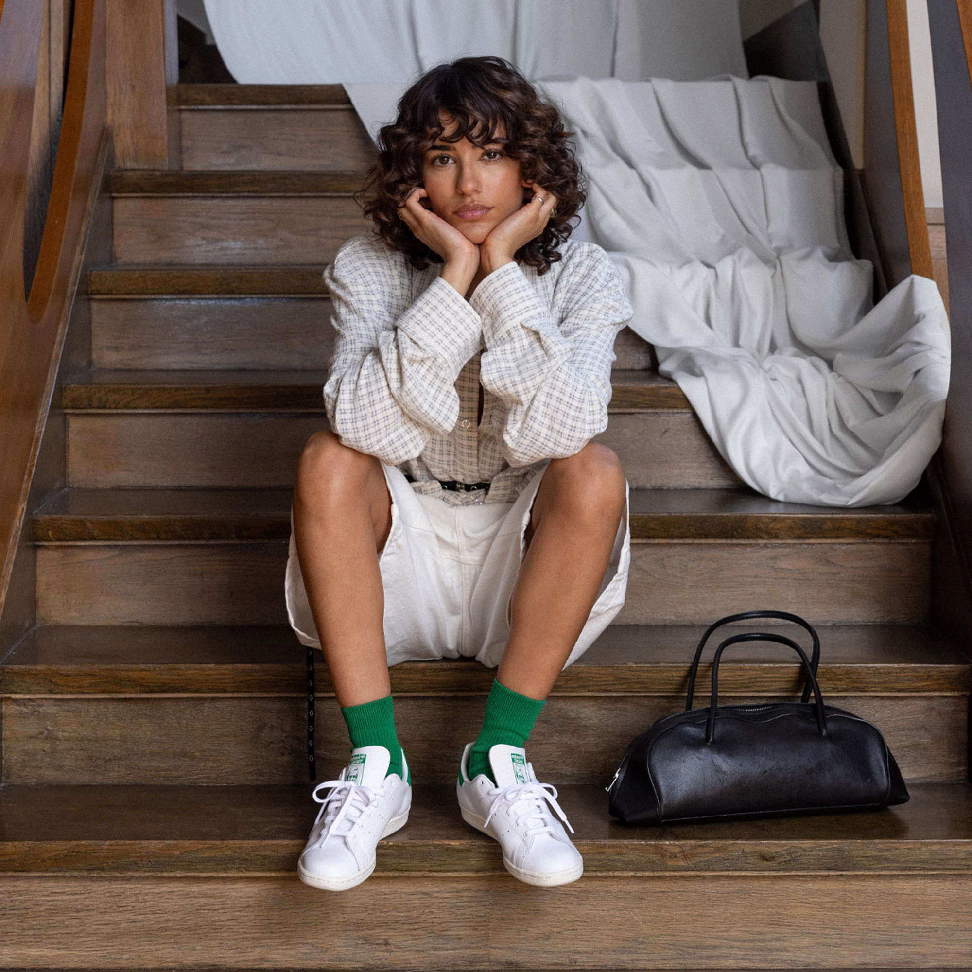 Person sitting on stairs, wearing a white outfit and green socks, with a black bag.