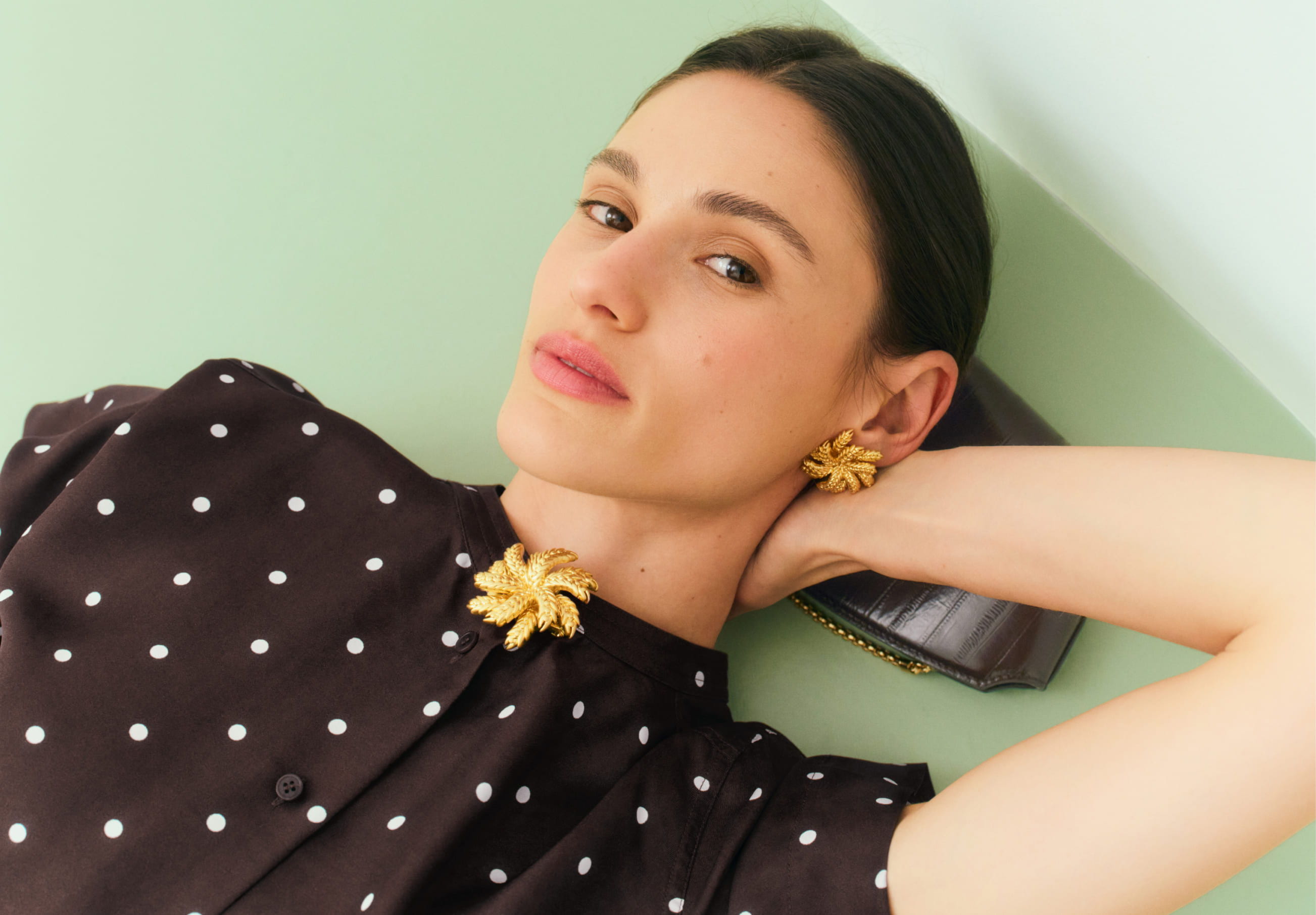 Person with dark hair wearing polka dot clothing and jewelry, resting against a wall.