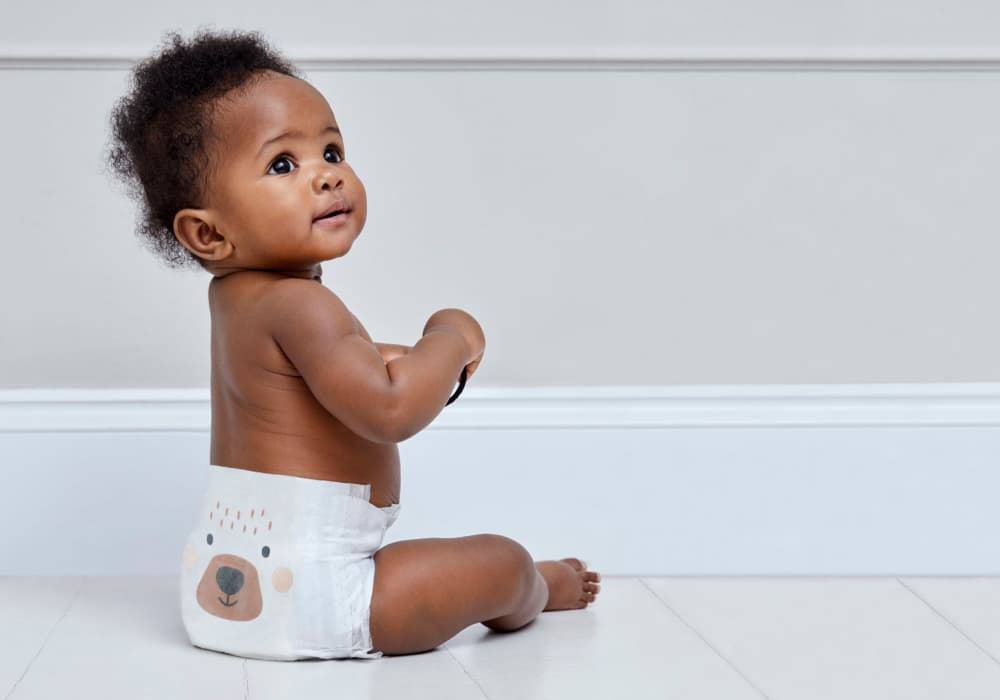 A baby sits on a white tiled floor wearing a Kit and Kin nappy with a bear print on the bottom.