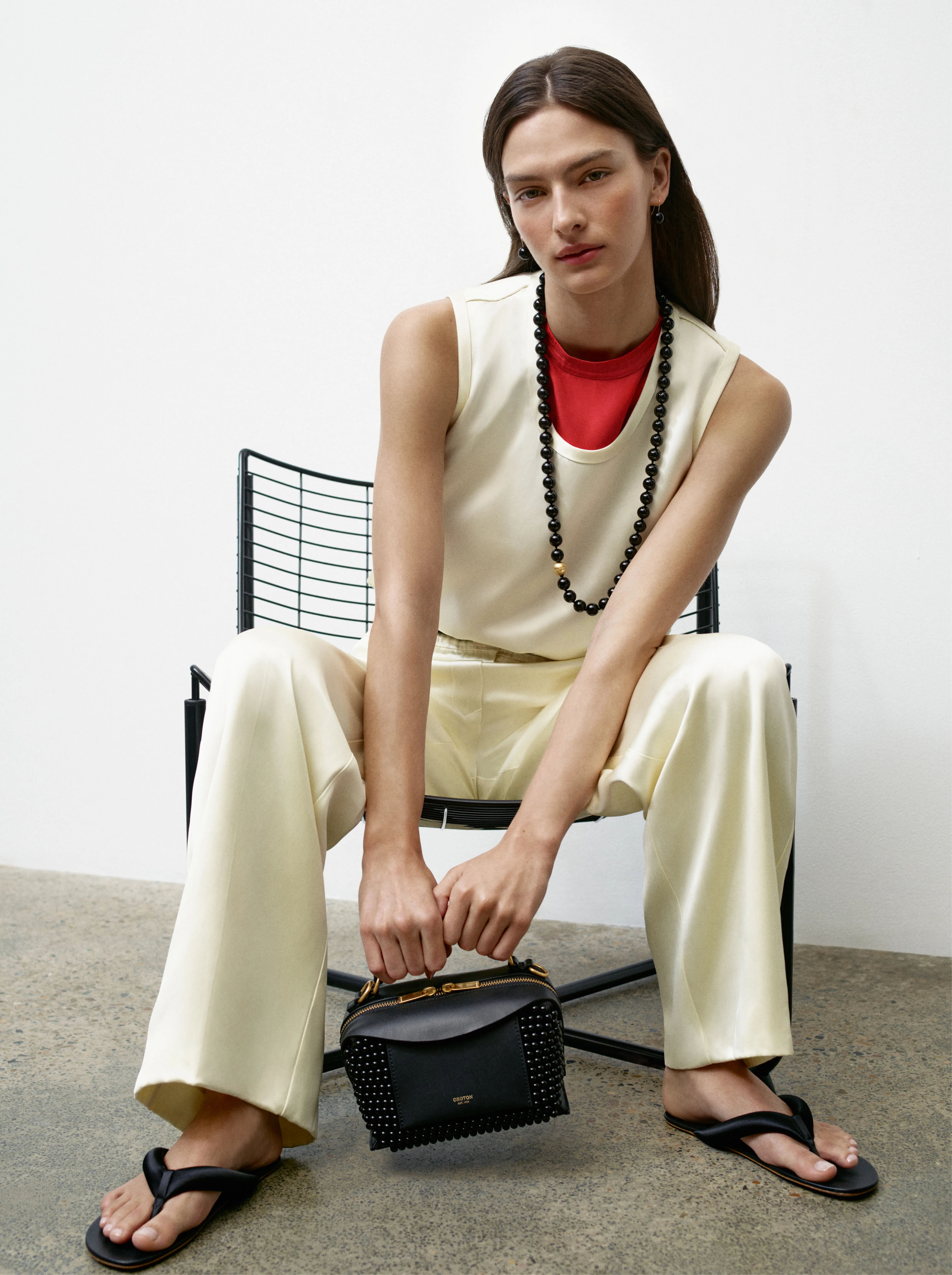 Model seated in a chair, wearing a stylish outfit and holding a handbag.