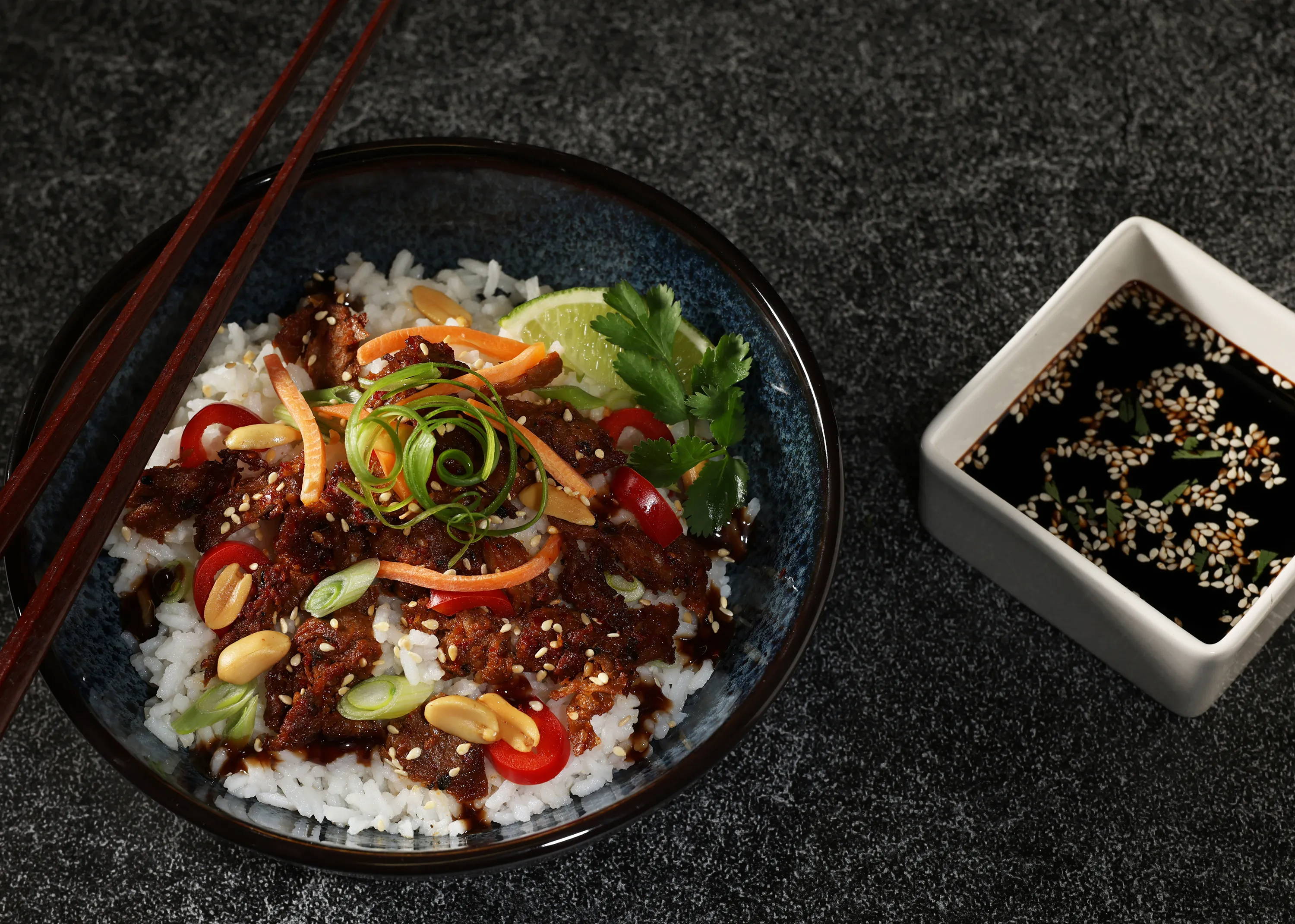 Harvest Shreds spiced with Bulgogi Blend on a bed of rice, with red peppers, scallions, peanuts, carrots and sesame seeds. In a large dark bowl with chopsticks and a square white ramekin with soy sauce and sesame seeds