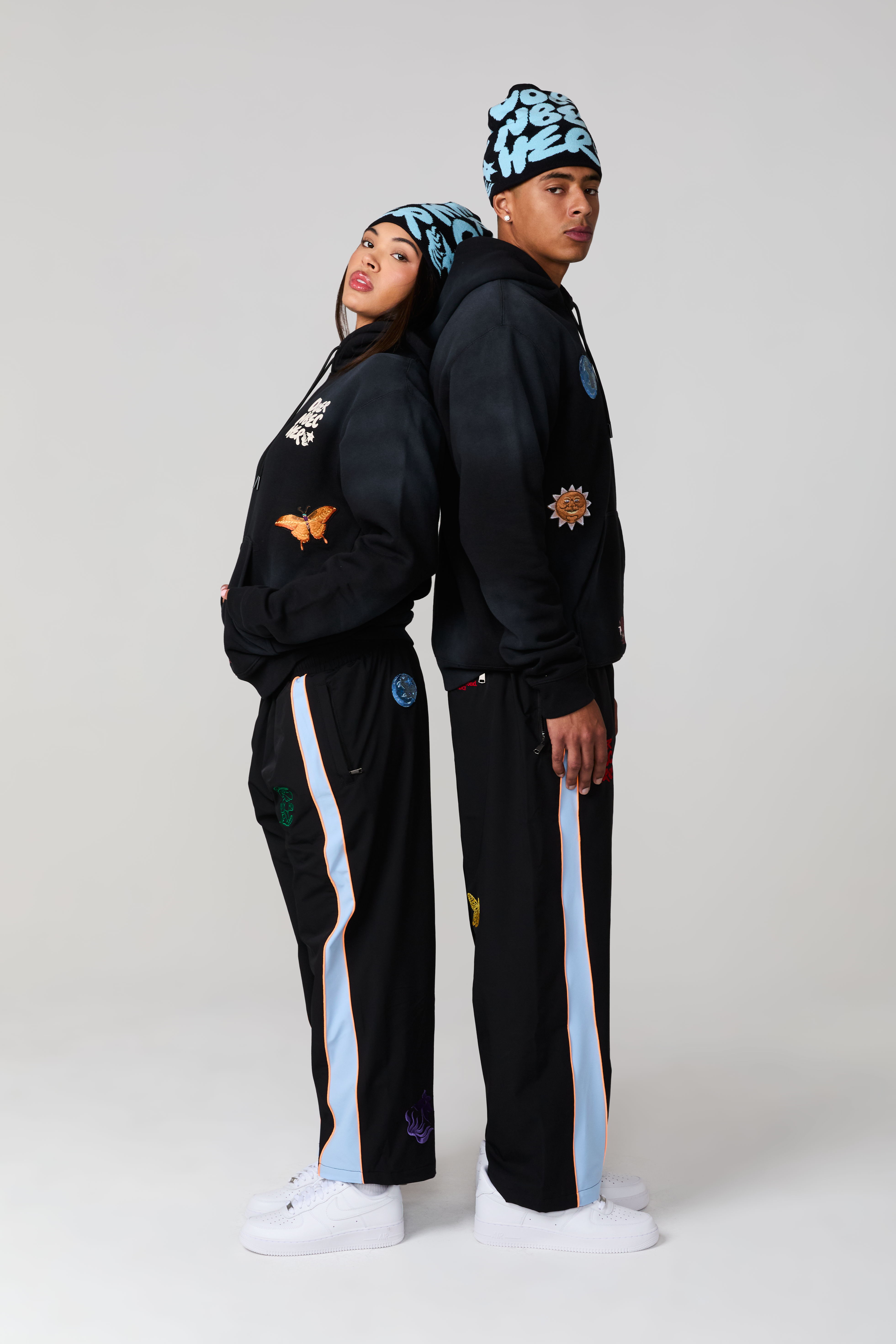 Two models in matching streetwear outfits, standing back to back.