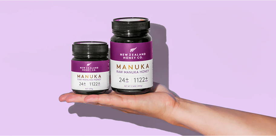 A hand holding two bottles of Manuka honey against a purple background.