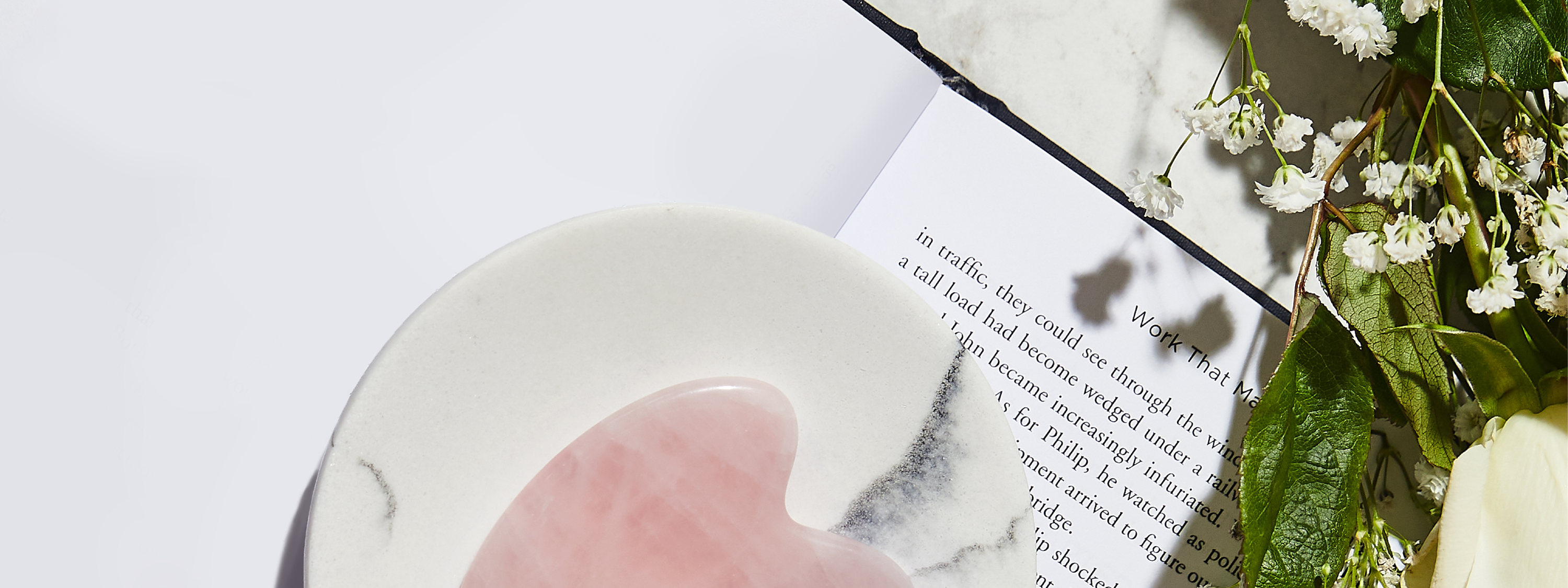 Open book holding light pink gua sha tool on top of small white and black marble plate surrounded by greenery and baby’s breath flowers.