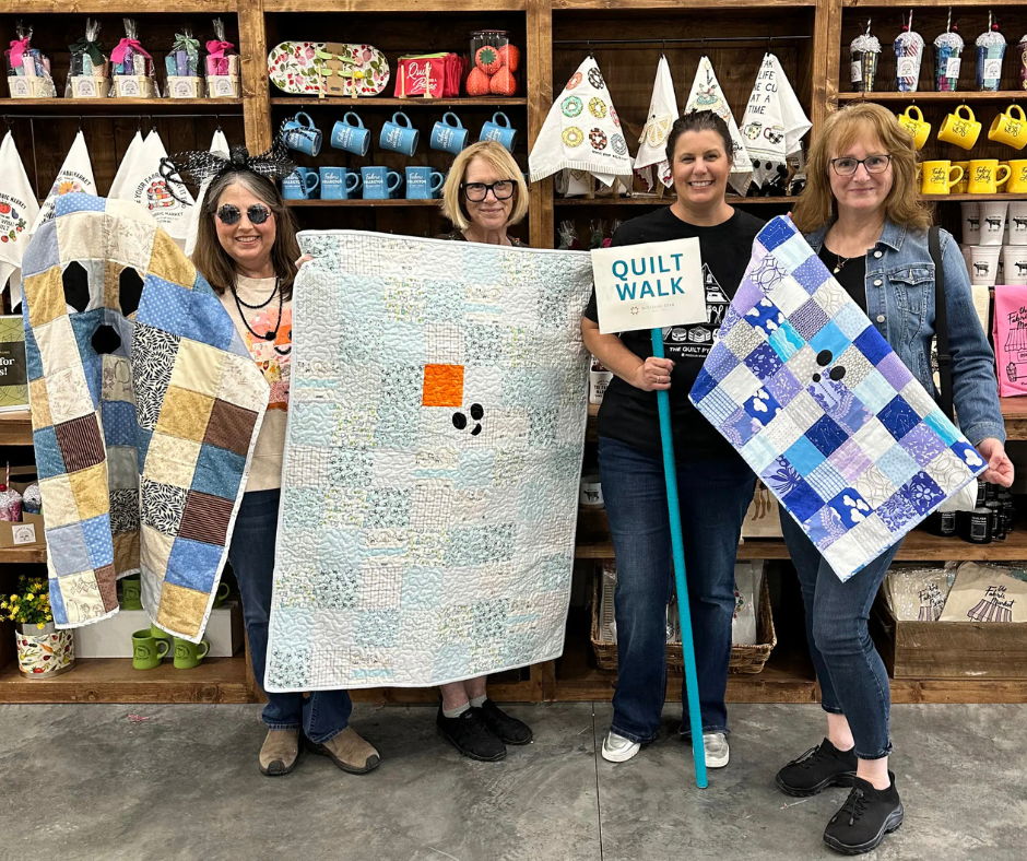 Four women holding quilts in a fabric store, smiling together.