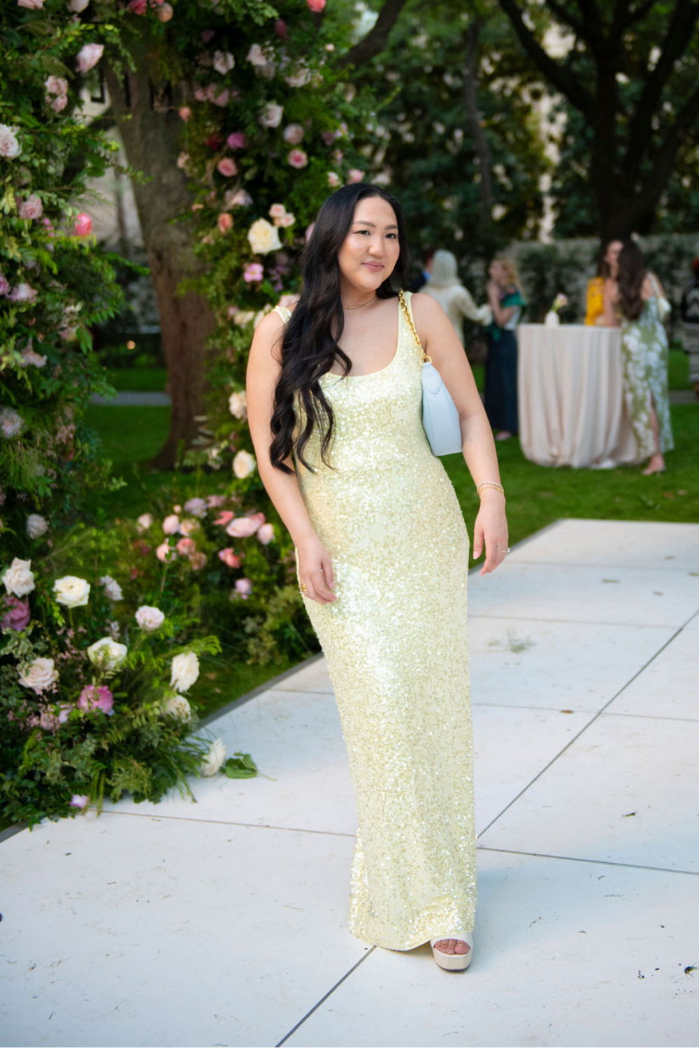 Woman in a sparkling yellow dress walking outdoors near floral decorations.