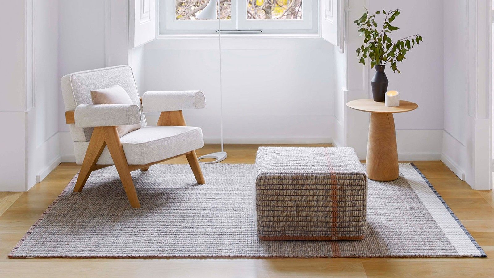 A cozy living room with a white chair, pouf, rug, and decorative plant.