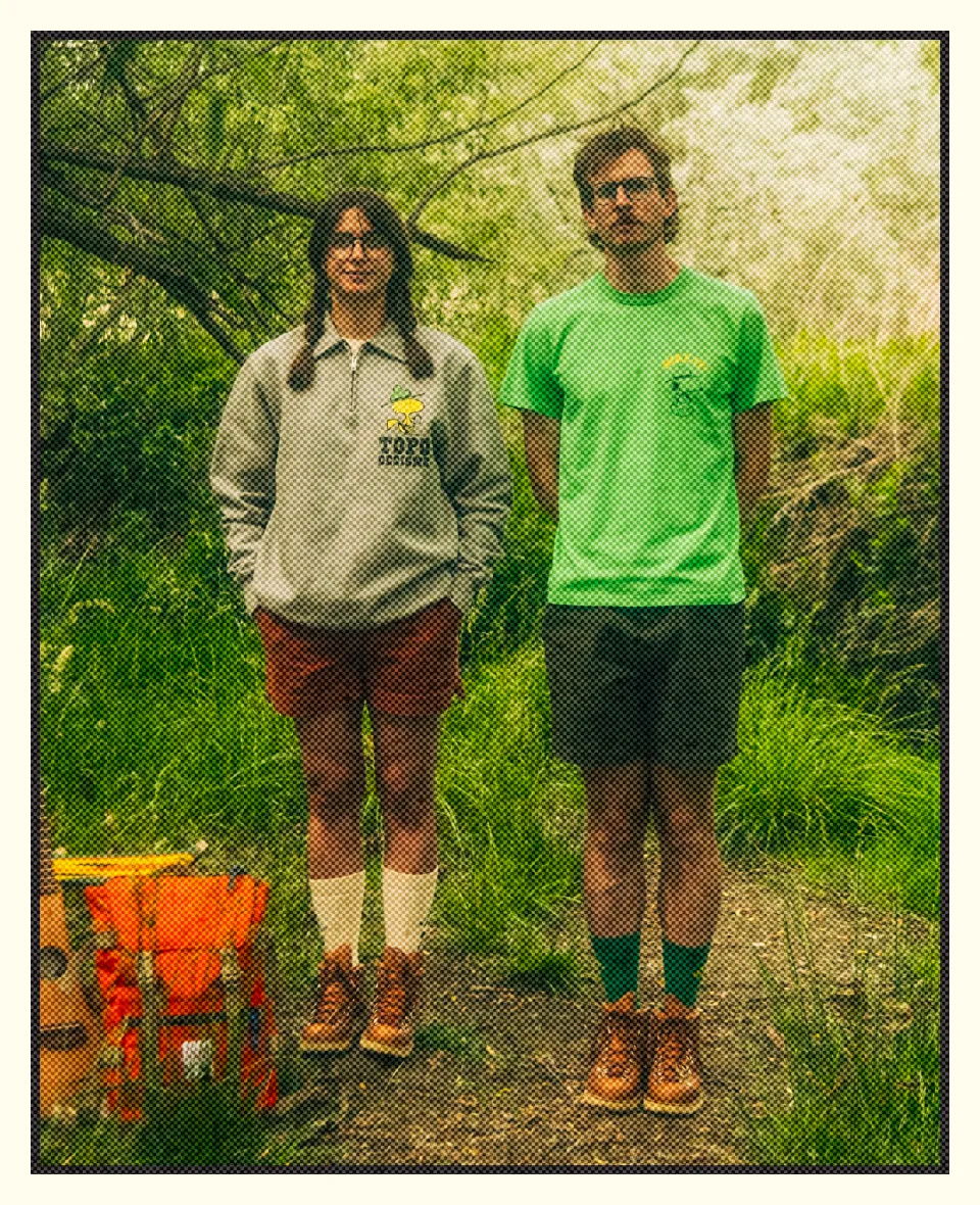 Two people standing in a forest, wearing casual clothing and boots.