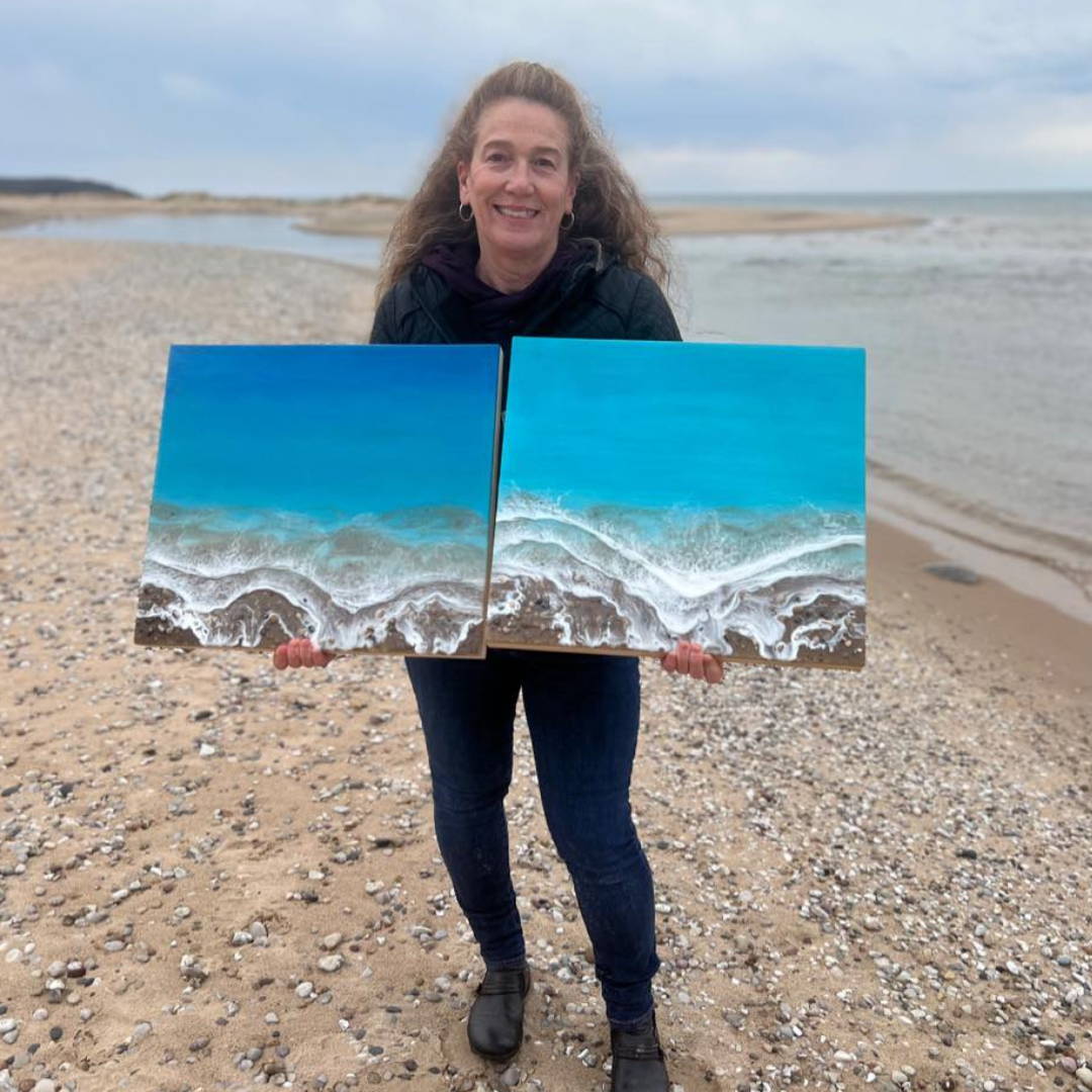 artist karen hammond standing on the beach holding two panels of ocean resin art she created