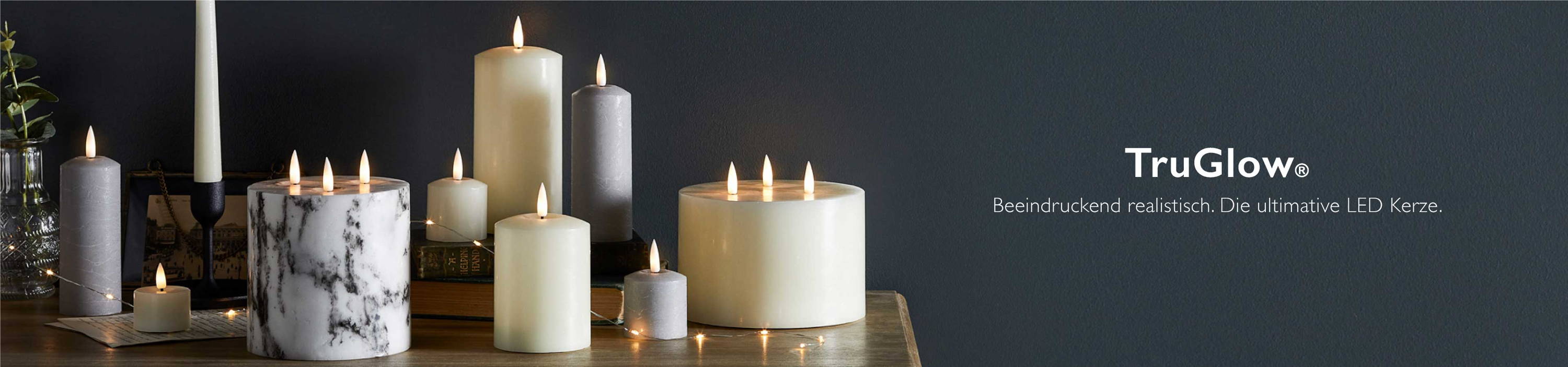 TruGlow LED Kerzen verschiedener Formen und Größen in Grau und Elfenbein auf einer Holzkommode vor einer dunklen Wand