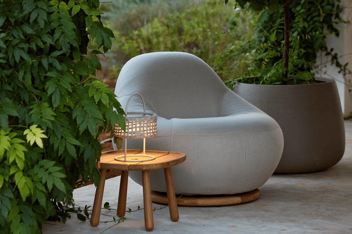 Cozy outdoor corner with a rounded gray lounge chair, woven lantern-style light on a small wood side table, and a large planter surrounded by greenery.