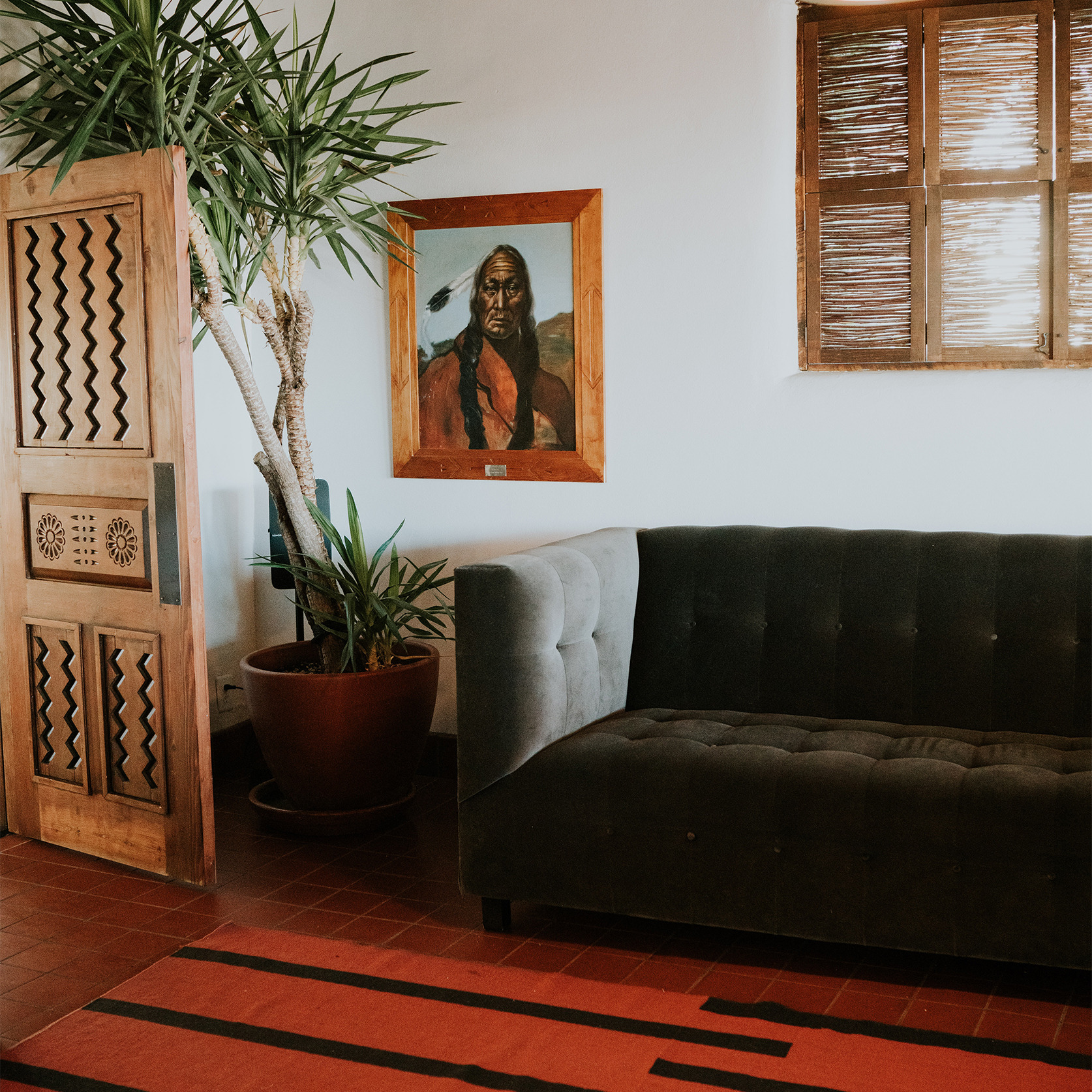 An ethnic furnished corner of a house with a painting of a Native American hanging on the wall, a beautifully carved wooden door and a sofa.