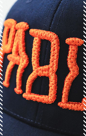 Close-up of navy cap with raised orange quilted puff embroidery of stylized lettering, showing the quilted texture created with stitching pattern