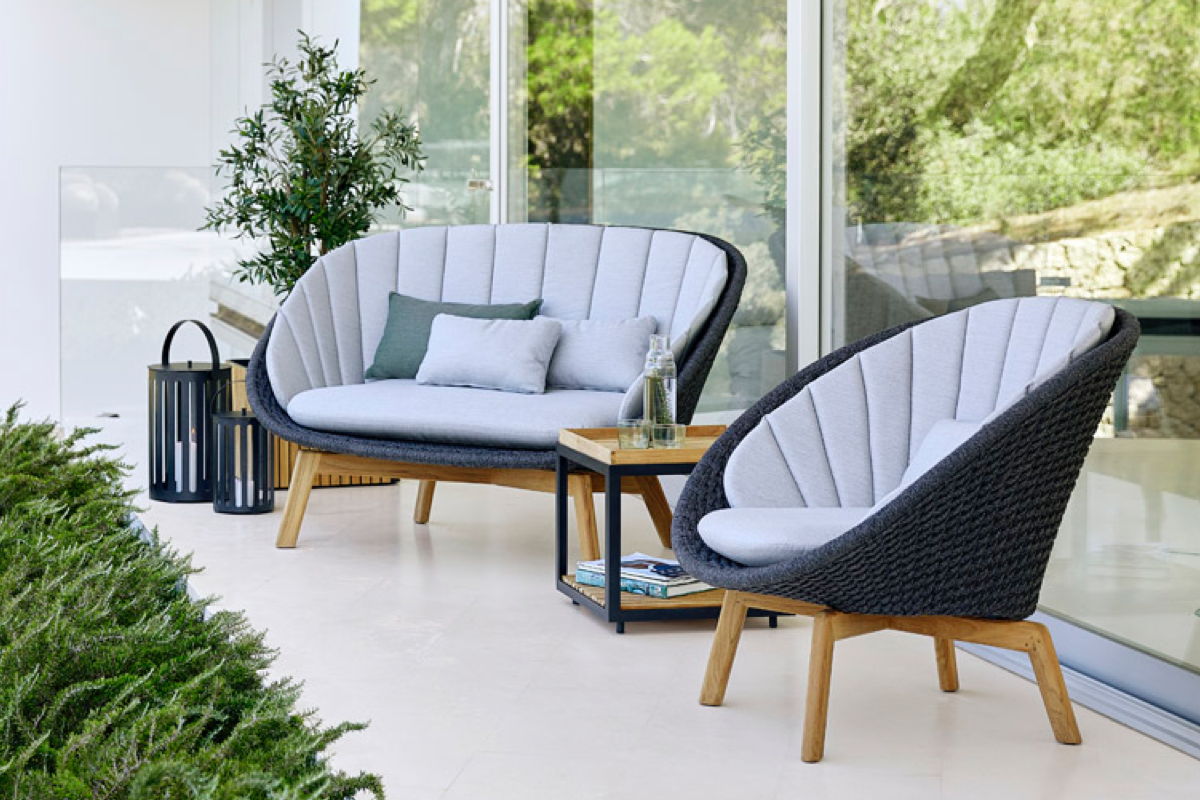 Outdoor lounge seating set with a woven loveseat and matching chair with light cushions and teak legs, paired with a small side table and black lanterns on a covered patio.