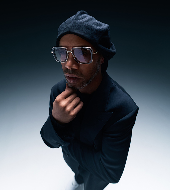 A person in sunglasses and a beanie posing thoughtfully against a dark background.