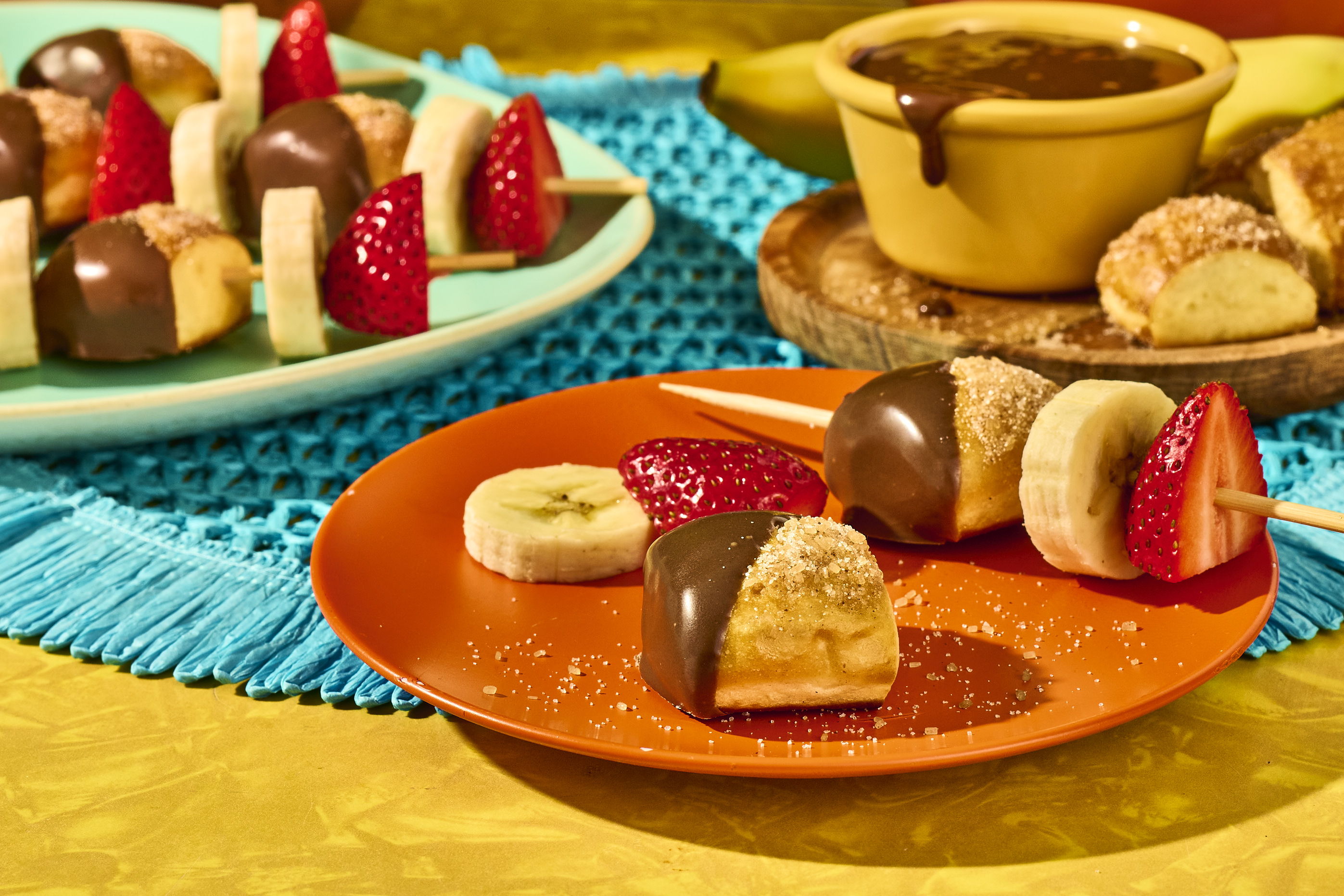 Chocolate-dipped fruit skewers with banana and strawberry on colorful plates.