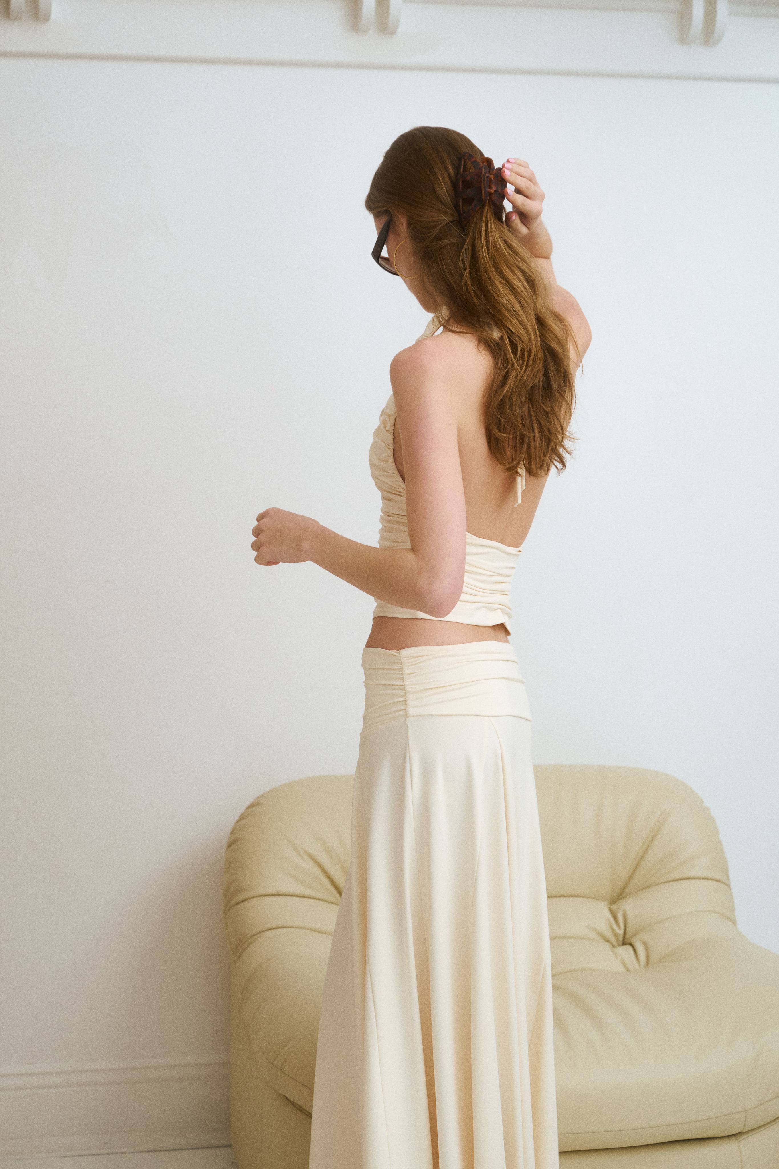 Woman in a beige dress adjusting her hair, standing near a couch.
