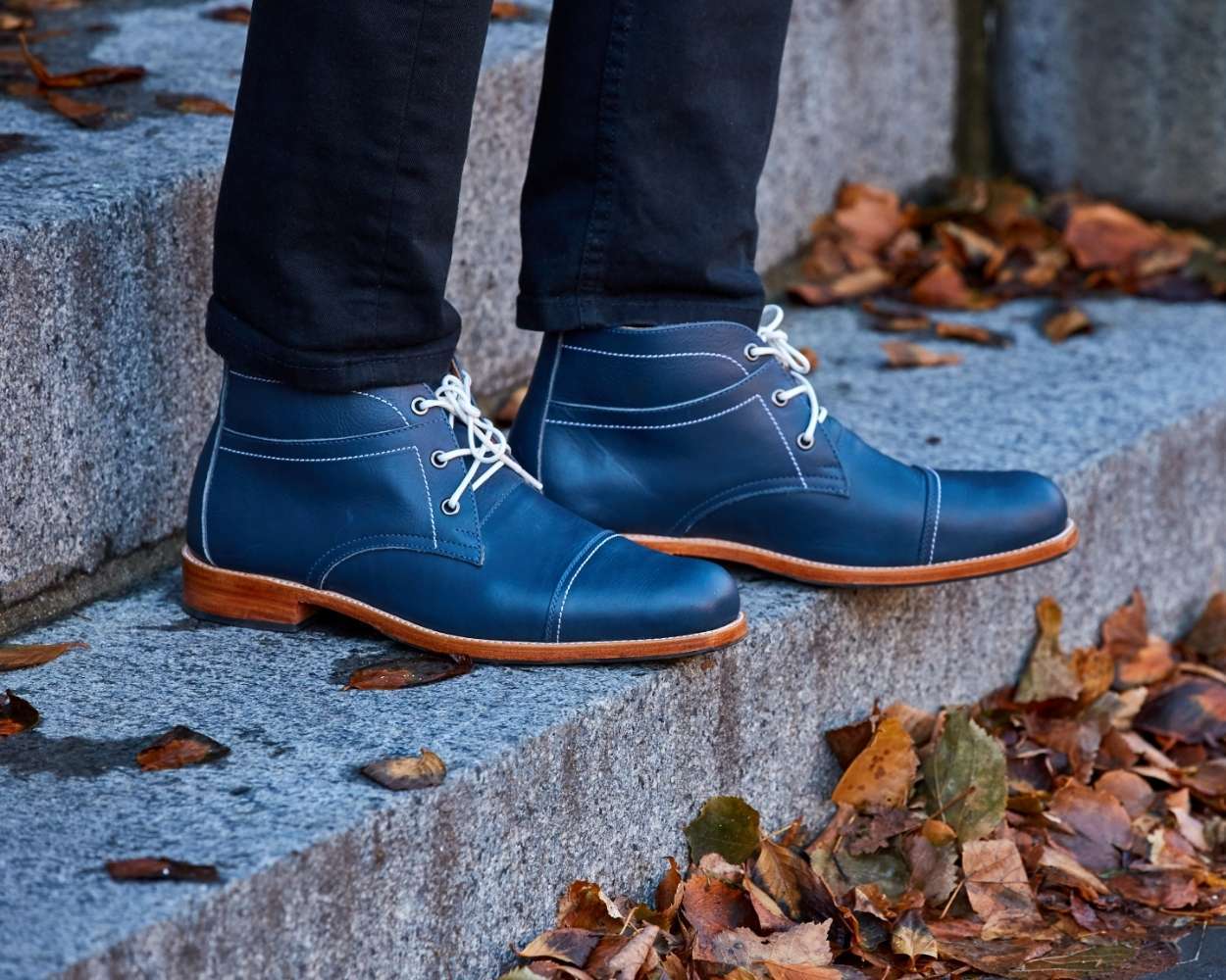 A close-up outdoor lifestyle shot of a man's feet standing on granite stone steps scattered with fallen autumn leaves. He is wearing denim blue leather Adelante Valparaiso chukka boots featuring prominent white contrast laces, white side stitching, and a light-colored stacked leather sole. The look is styled with black slim-fit pants.
