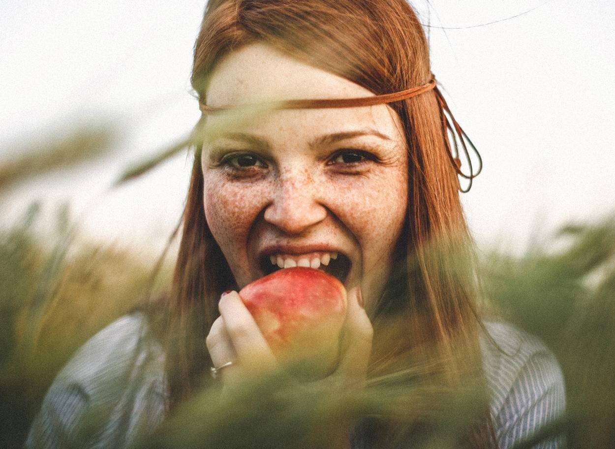 Šťastná dlouhovlasá mladá žena venku kouše do červeného jablka, ze kterého by může mít mravenčení v ústech, pokud má alergii na pyl břízy