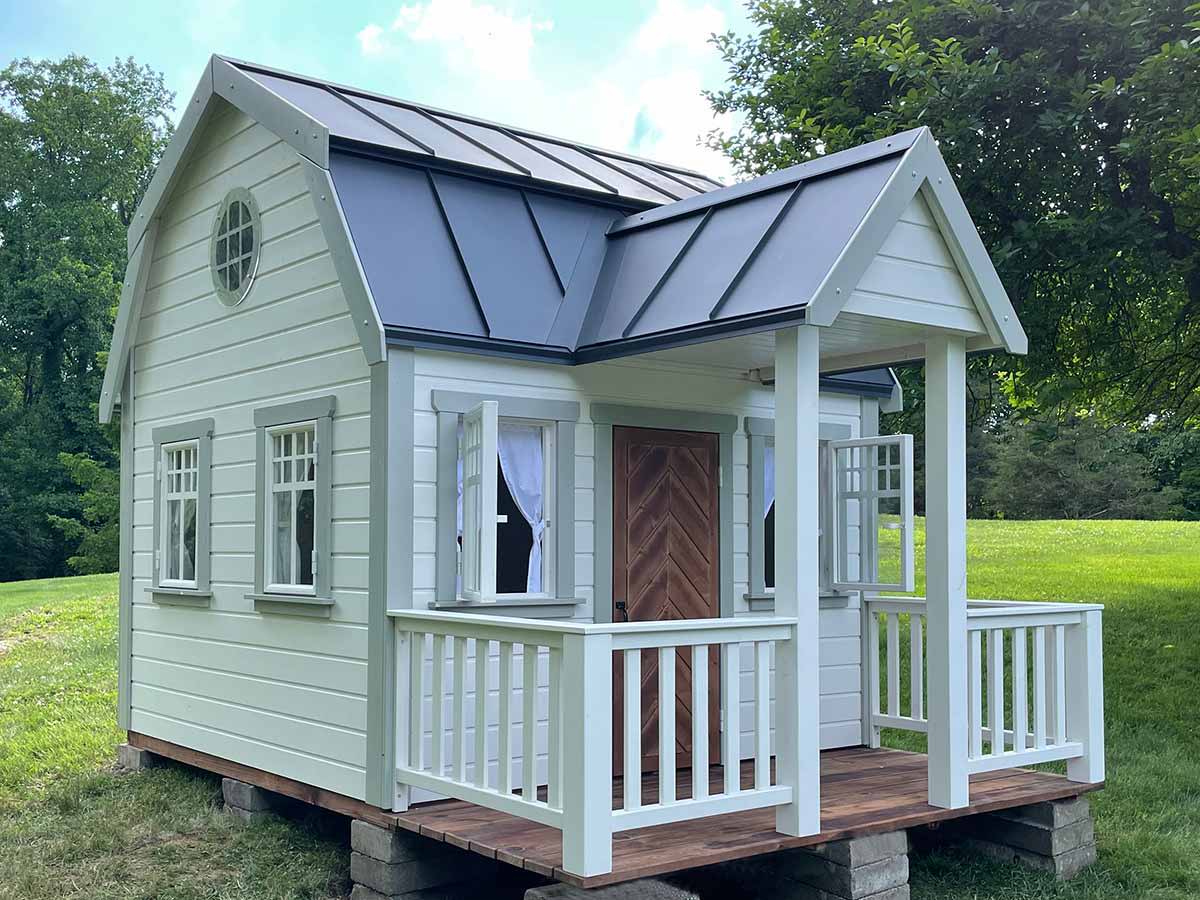 Farmhouse Style Outdoor Playhouse with porch, grey metal roof and the round top window in a backyard by WholeWoodPlayhouses
