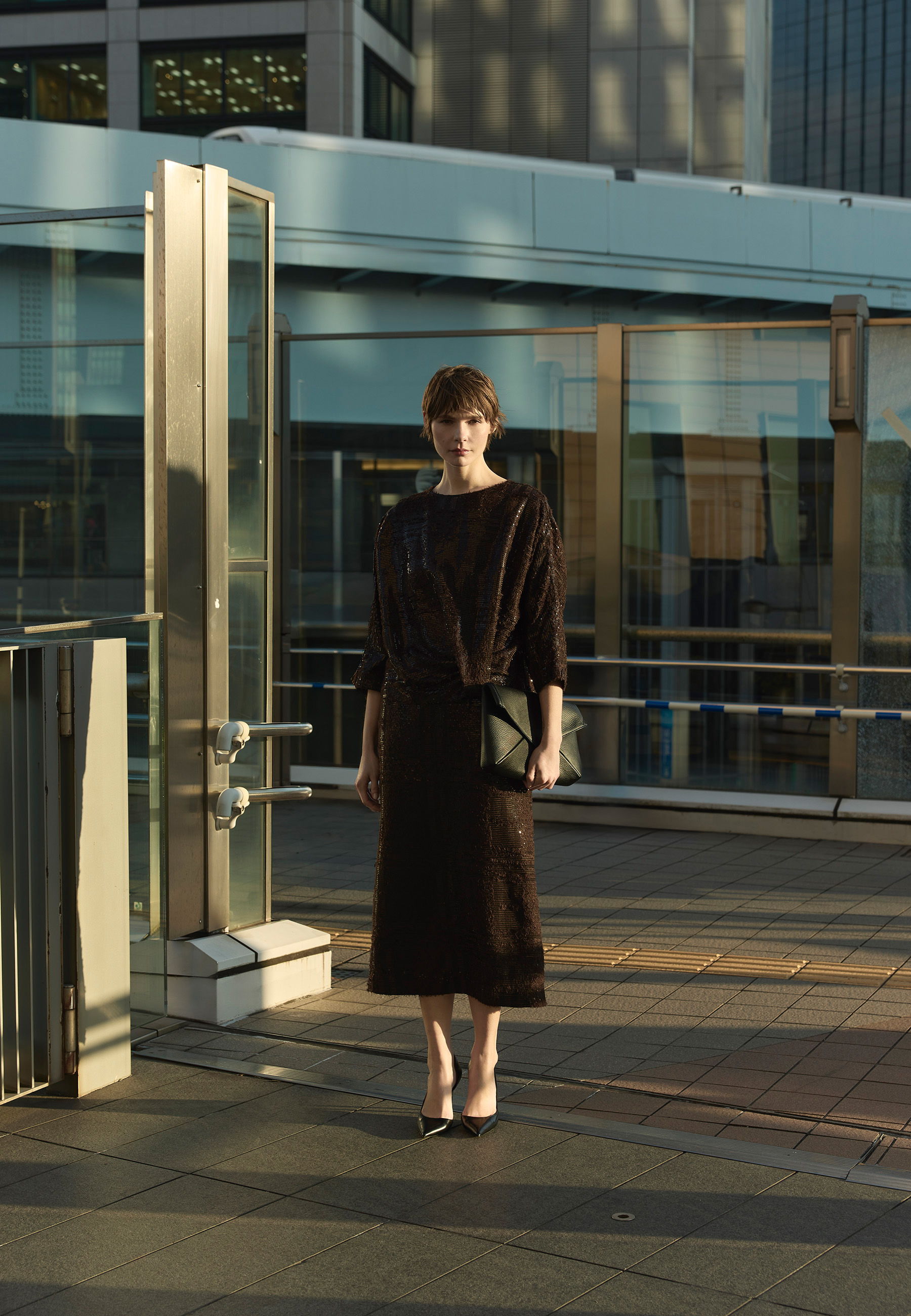A woman in a black dress stands outdoors near modern architecture.