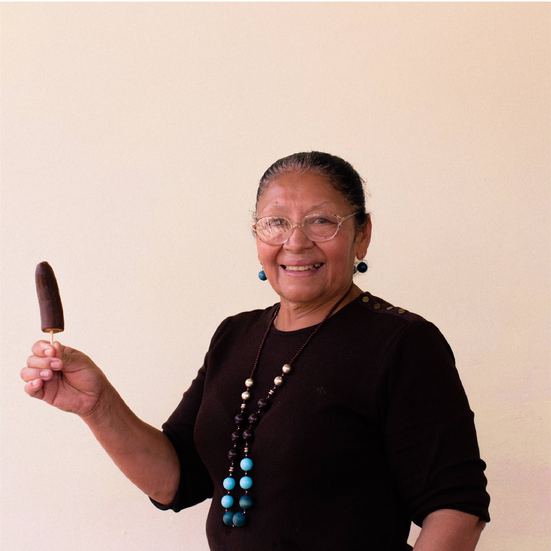 A Honduran woman stands and smiles while holding up her homemade chocolate covered banana on a stick.