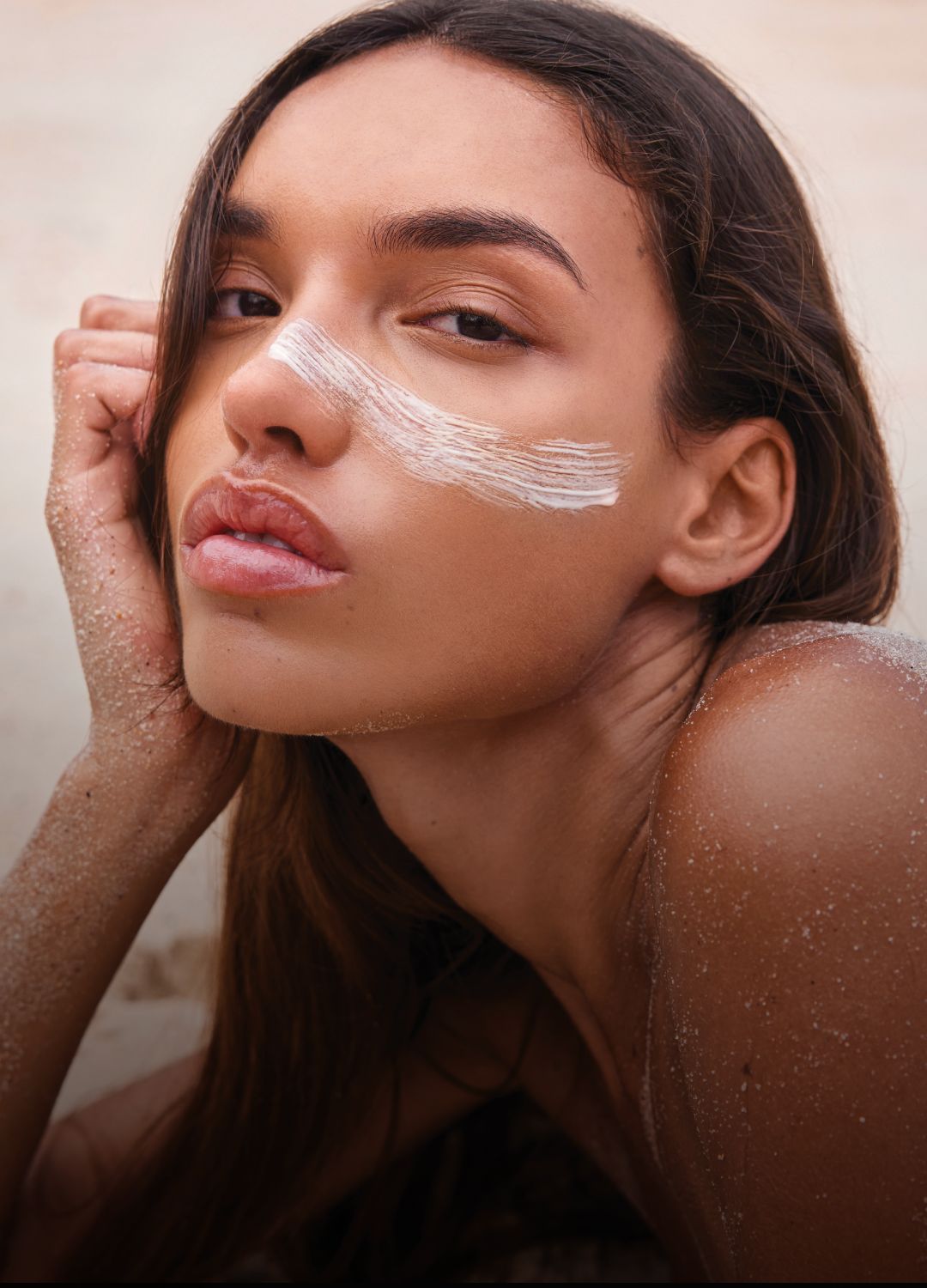 Woman with facial cream lying on sandy surface, gazing at the camera.