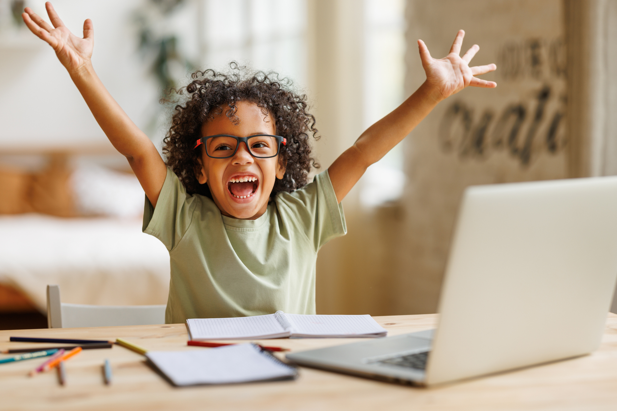 Child celebrating with arms raised, sitting at a desk with a laptop.
