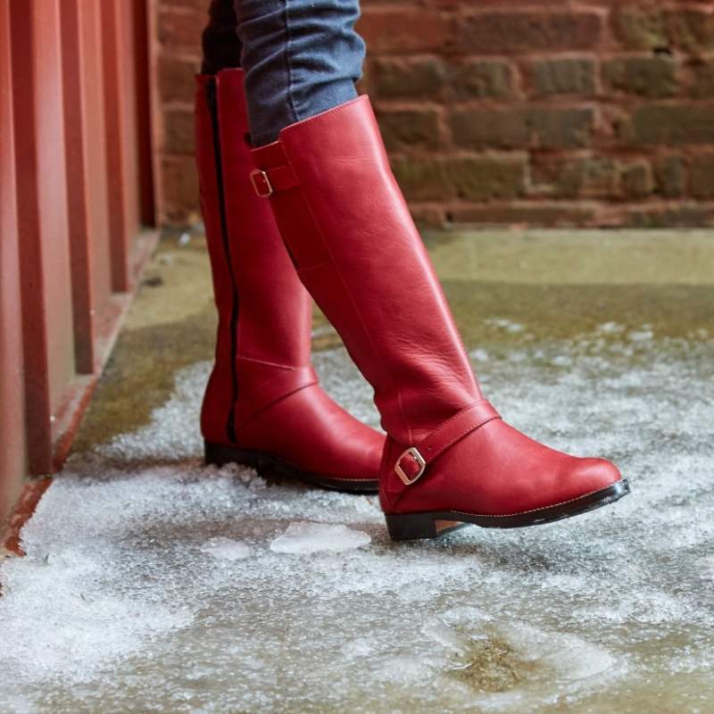 Woman wearing pomegranate leather Reina wide calf equestrian boots with an ankle strap buckle detail, standing on a snowy ground layout.