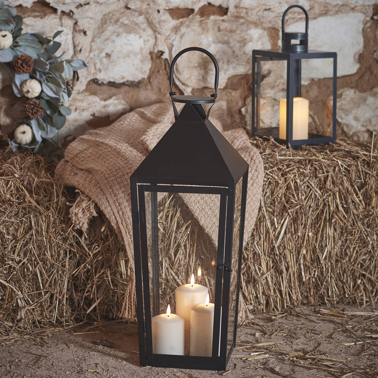 A black lantern with 3 LED candles inside and a smaller black candle lantern in the background in an autumn barn.
