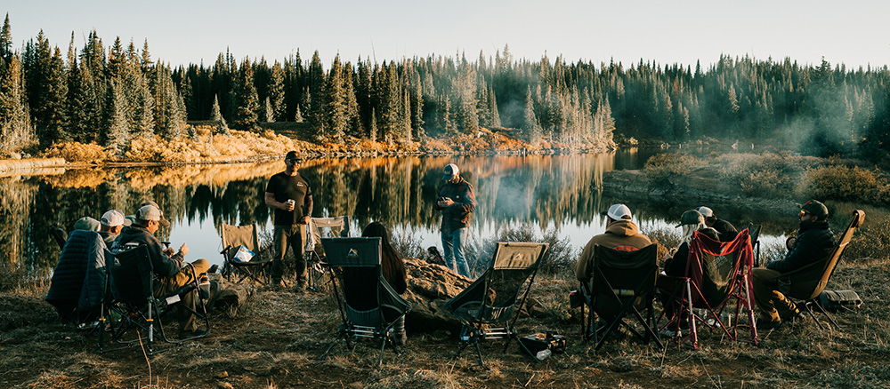 Group of people camping by a lake with a campfire in the background.