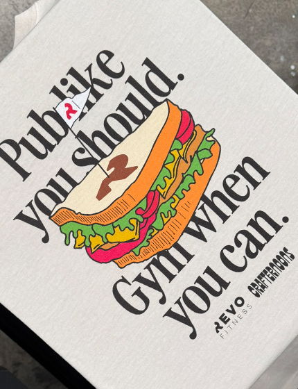 Minimalist typography t-shirt reading “Pub like you should. Gym when you can.” with sandwich graphic in bright tones.