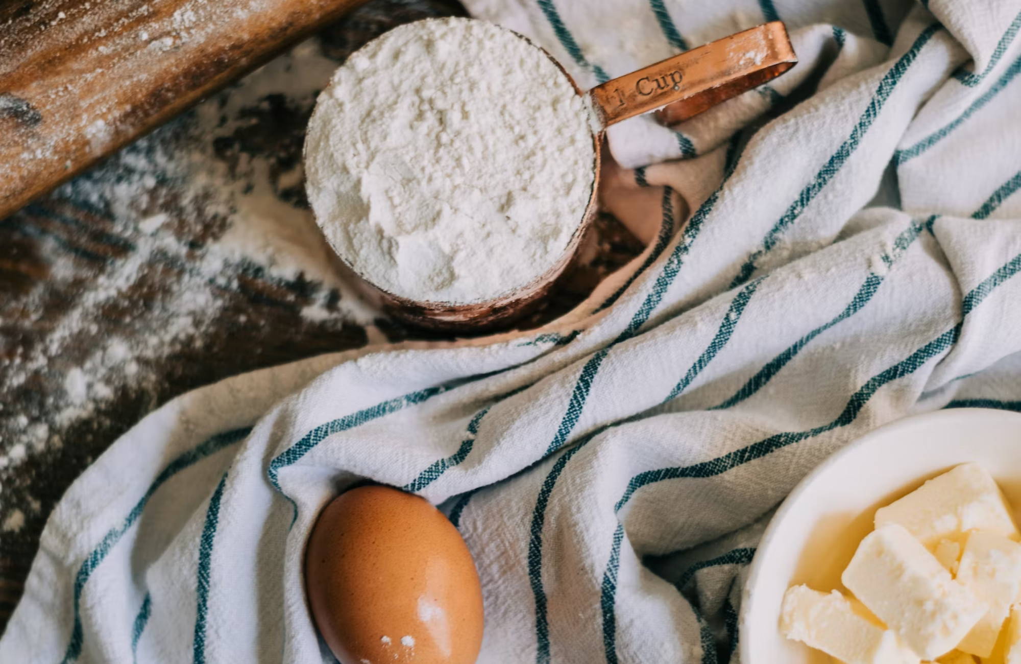 a cup of flour sitting on a table next to a blue and white towel, egg, butter, and rolling pin
