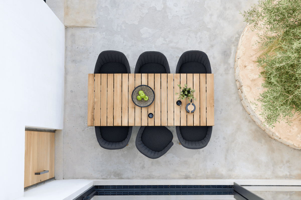 Top-down view of a rectangular teak outdoor dining table with black upholstered chairs on a minimalist concrete patio beside a planted bed. Caption (optional): Clean lines, warm wood, and deep seating make this outdoor dining area feel simple, grounded, and ready to use.