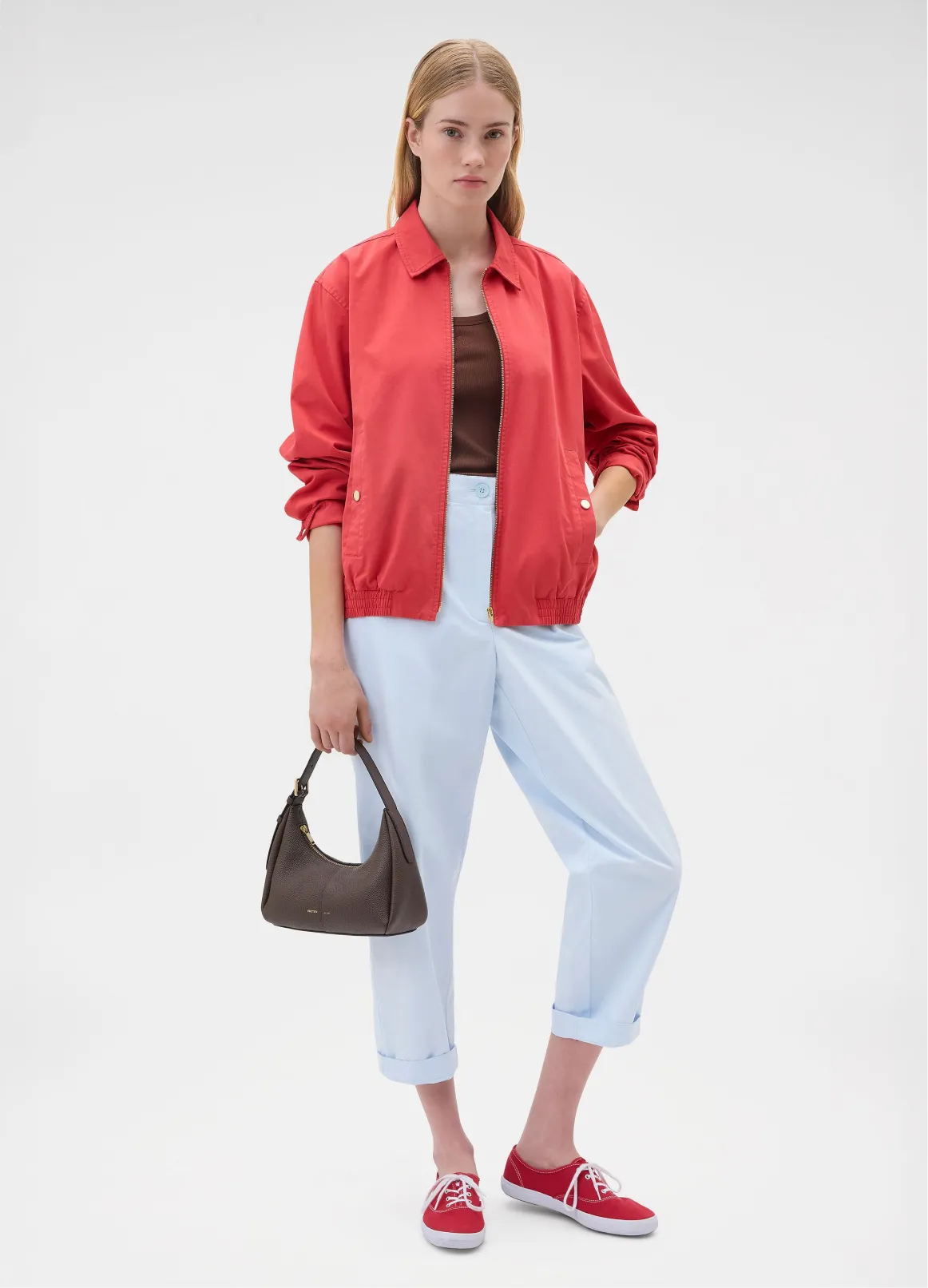 Woman in red jacket, brown top, light blue pants, and red shoes, holding a bag.