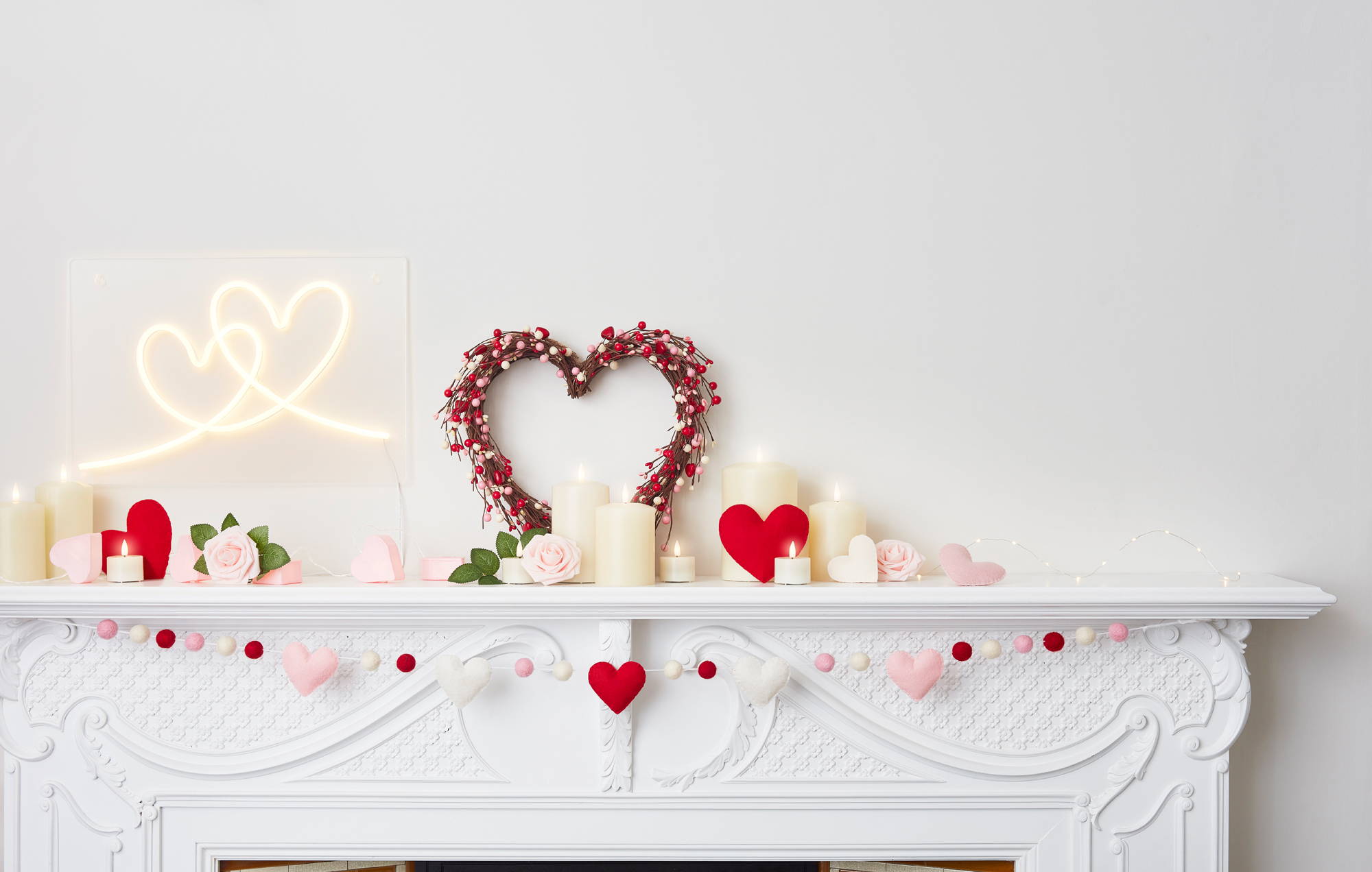 A white mantlepiece with a heart wreath, wall light, garland, felt decorations and candles.