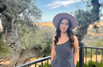 Woman in a gray dress and hat stands outdoors with trees and hills behind her.