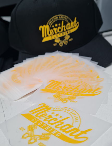 Black Cap with a yellow Merchant logo printed in ornate script sitting behind a pile of cut dtf film
