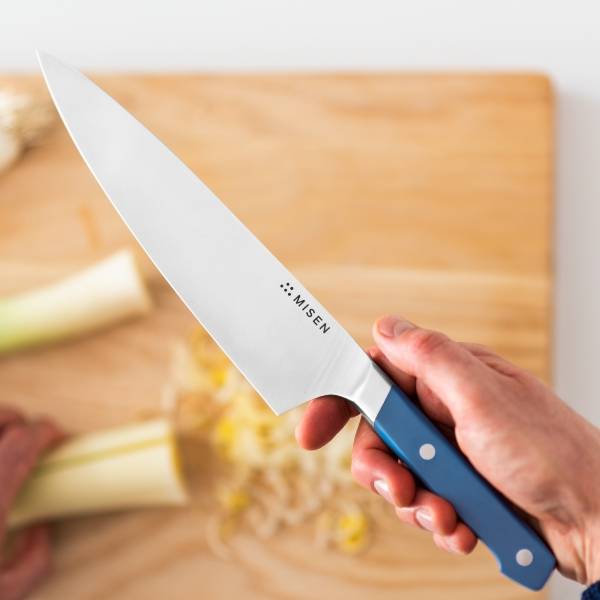 A hand holds a blue Misen Chef’s Knife with the blade’s flat side facing an overhead camera. Sliced leek on a light wood cutting board is visible in the background.