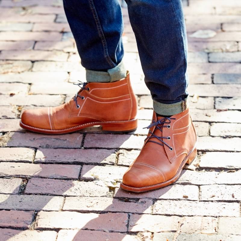 An outdoor lifestyle shot from the shins down of a person standing on a sunlit brick walkway. They are wearing a pair of light brown or caramel leather Adelante Valparaiso mid-top lace-up boots featuring dark laces, distinct contrast side stitching, a cap toe design, and a stacked leather sole. The look is styled with dark wash denim jeans with rolled cuffs.