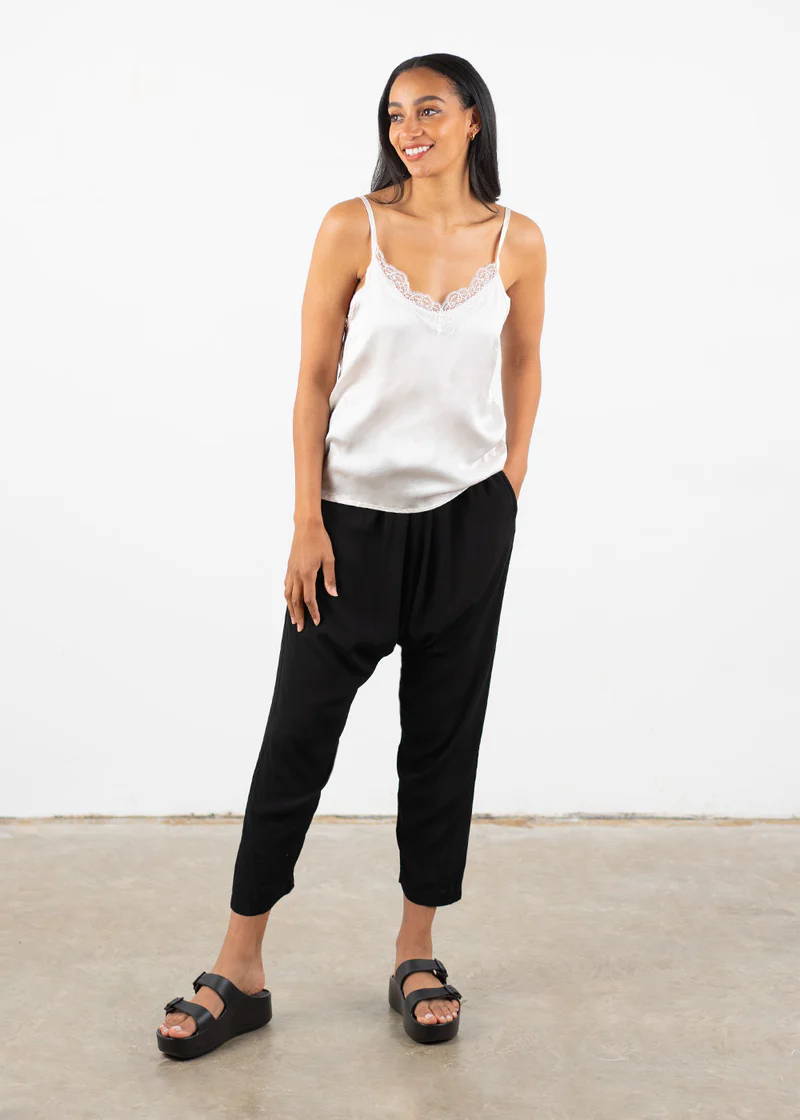 A model wearing an off white, silky camisole with lace detailing around the necklace, black cropped trousers and black chunky platform slides