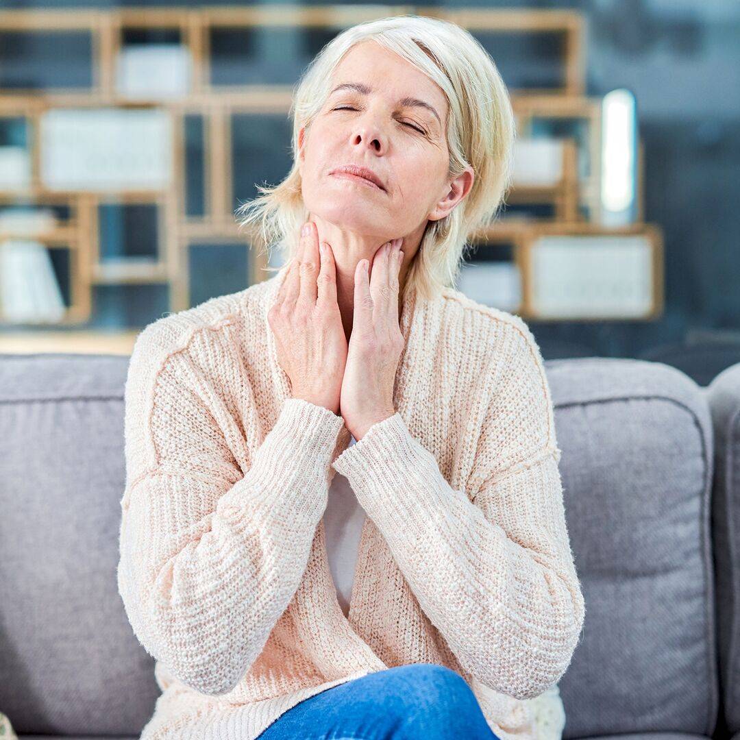 Žena sedící na pohovce, se zavřenýma očima a rukama dotýkajícími se krku. Možná její bolest v krku souvisí s alergiemi.