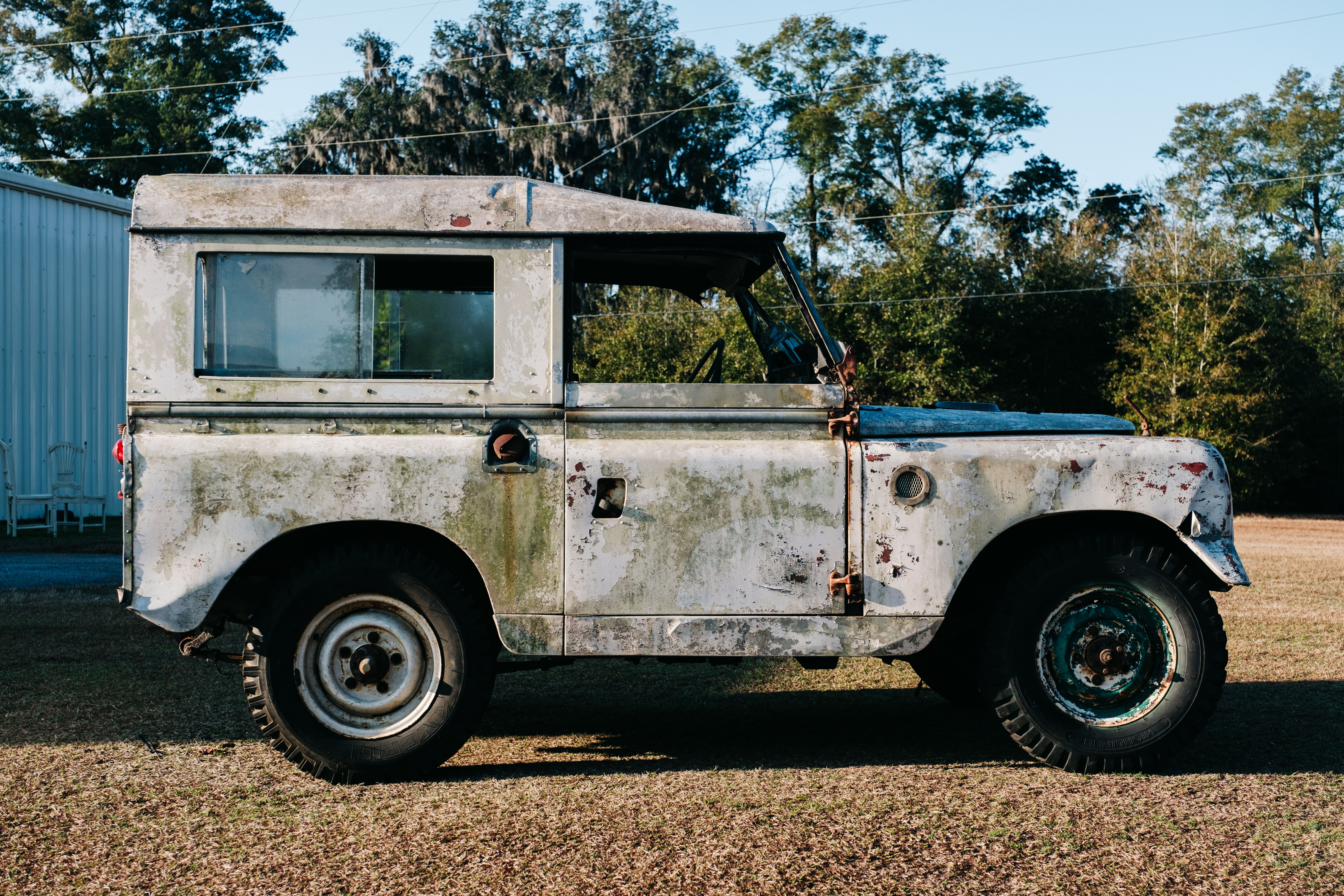 Weathered vintage vehicle parked on grassy area with trees in background.