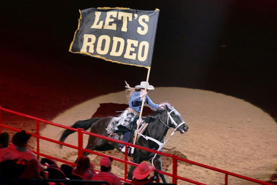 Cowgirl riding in the San Antonio arena with a LET'S RODEO flag.