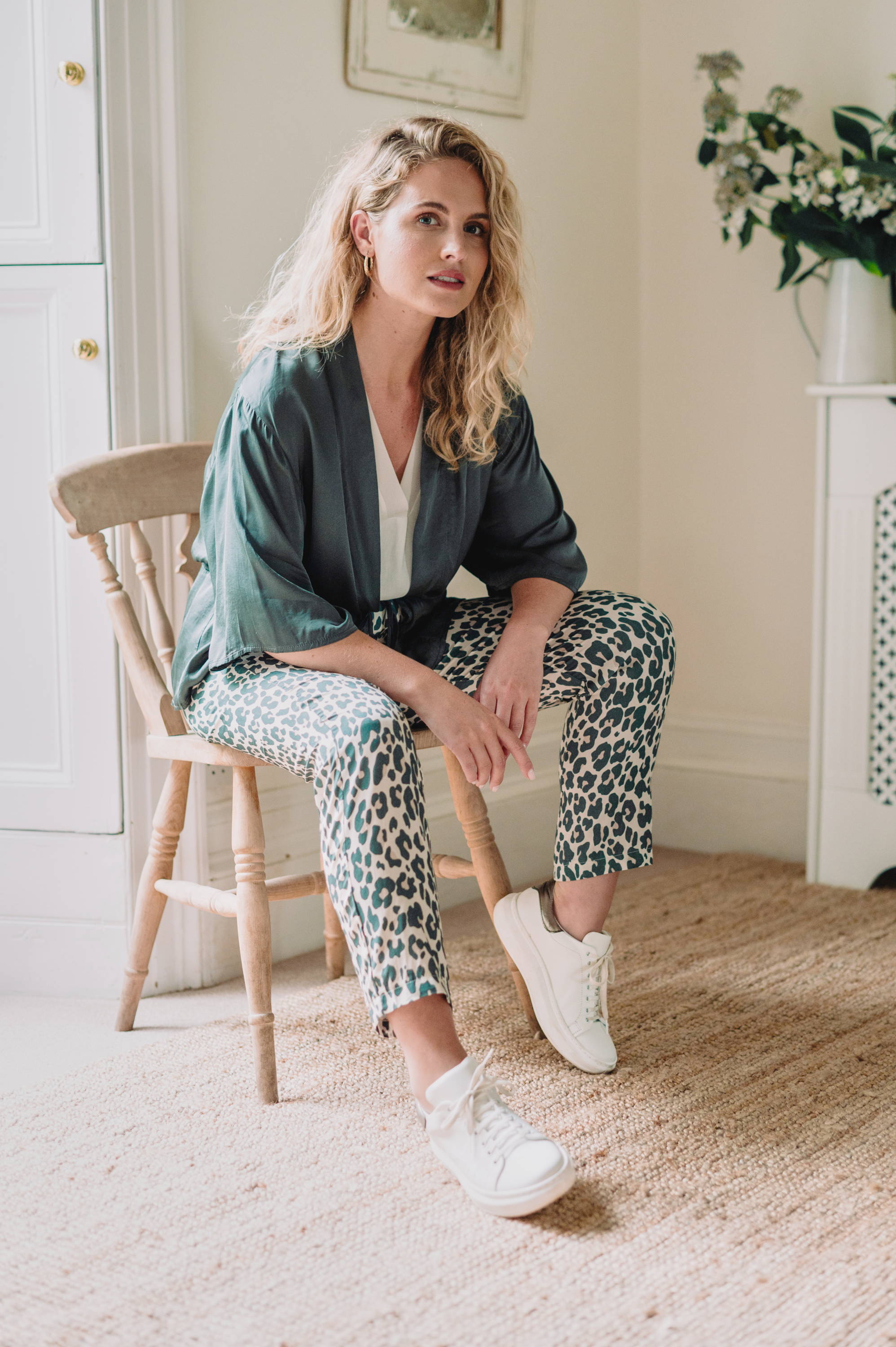 A model sitting on a woodne chair wearing a pair of leopard print, tapered trousers white, white top, dark grey satin jacket and white trousers 