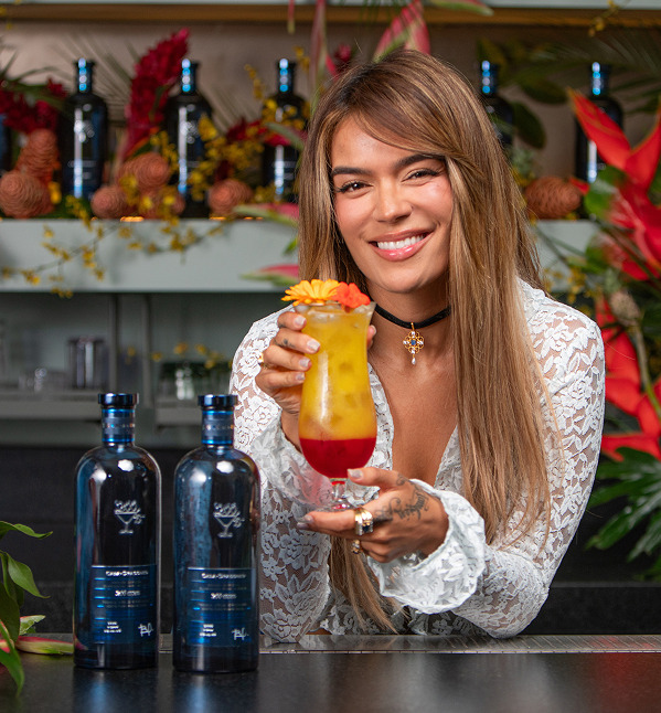 A woman smiles while holding a colorful cocktail, with bottles in the background.