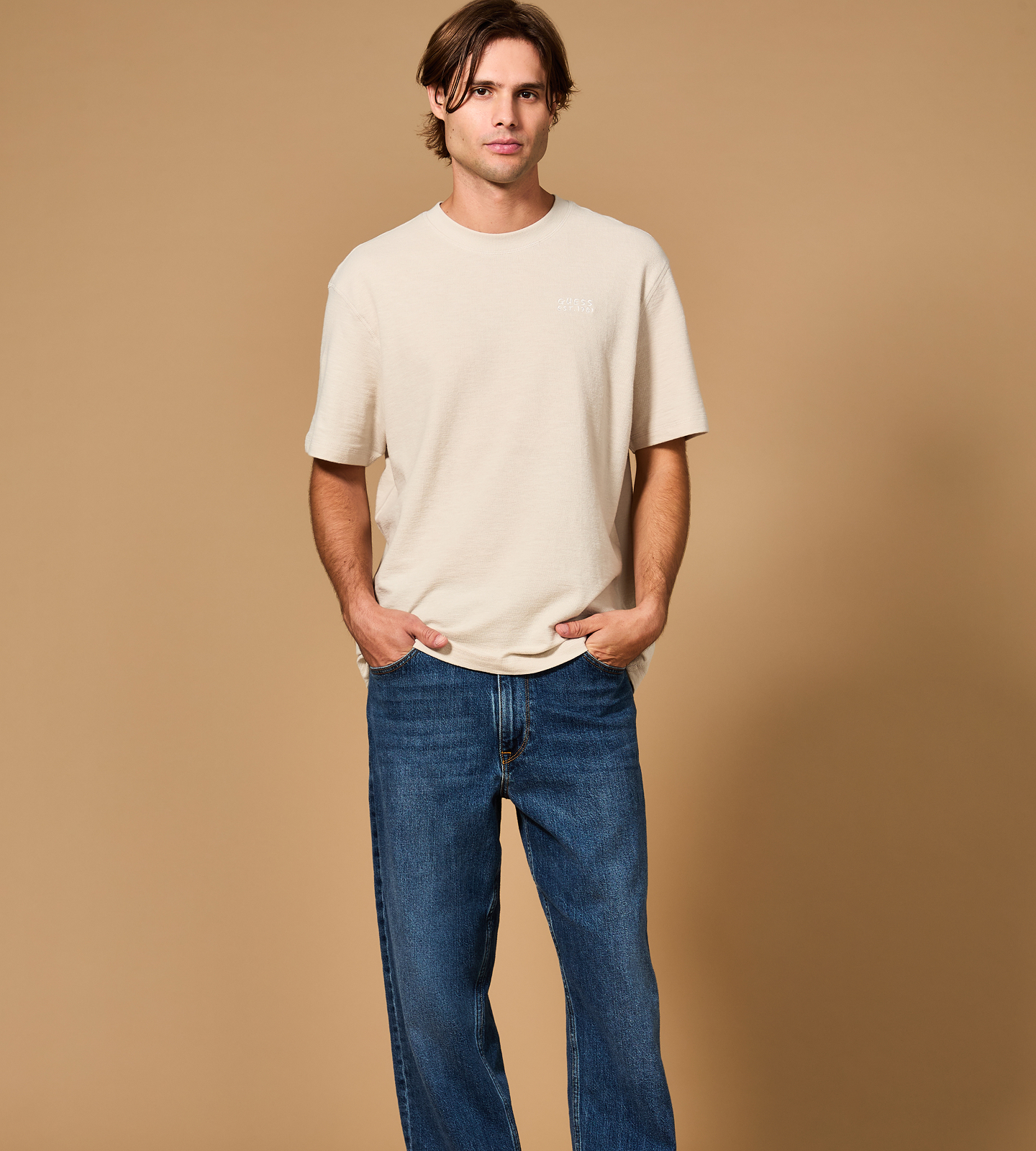 A man in a light shirt and blue jeans against a beige background.