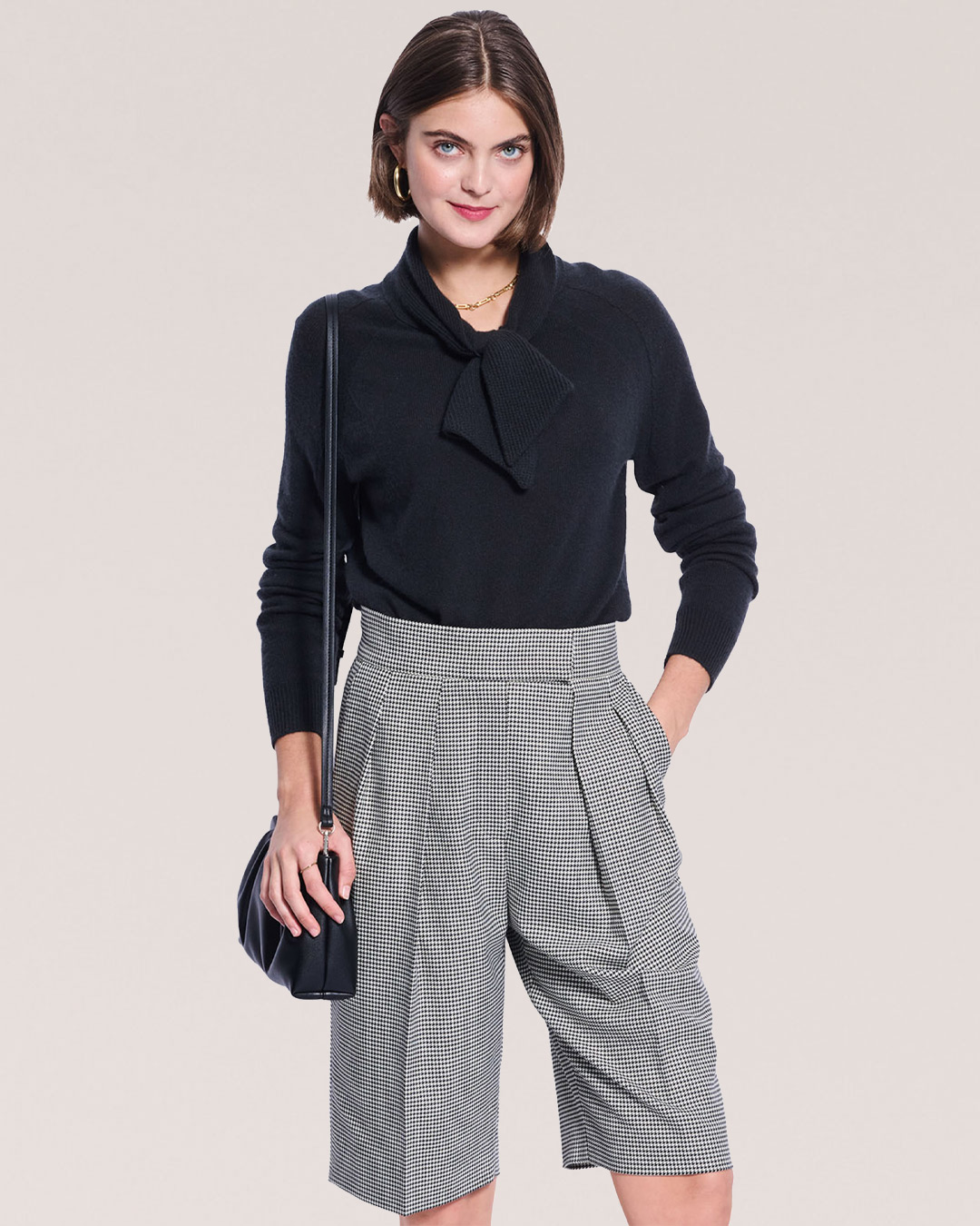 Woman in a black sweater and gray pleated shorts, standing confidently.
