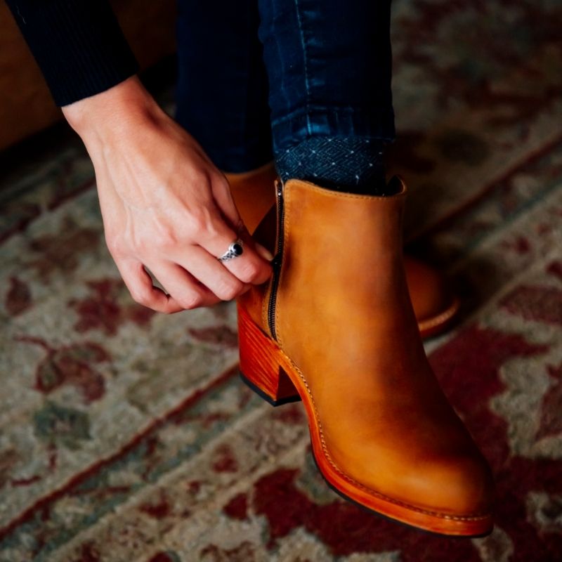 Close-up lifestyle view of a woman wearing the handcrafted Adelante Granada leather ankle boots in warm caramel brown, adjusting her boot while standing on a patterned vintage rug.