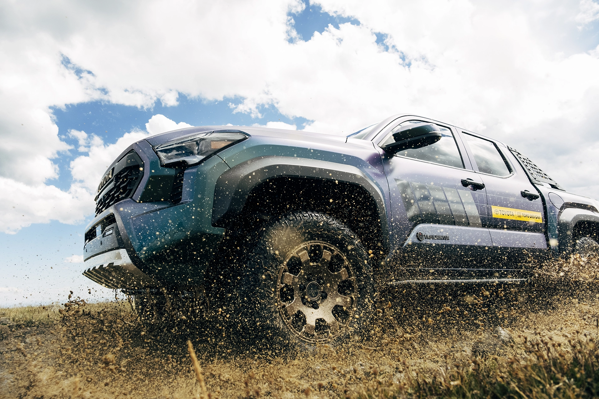 A blue truck driving through a muddy landscape under a cloudy sky.